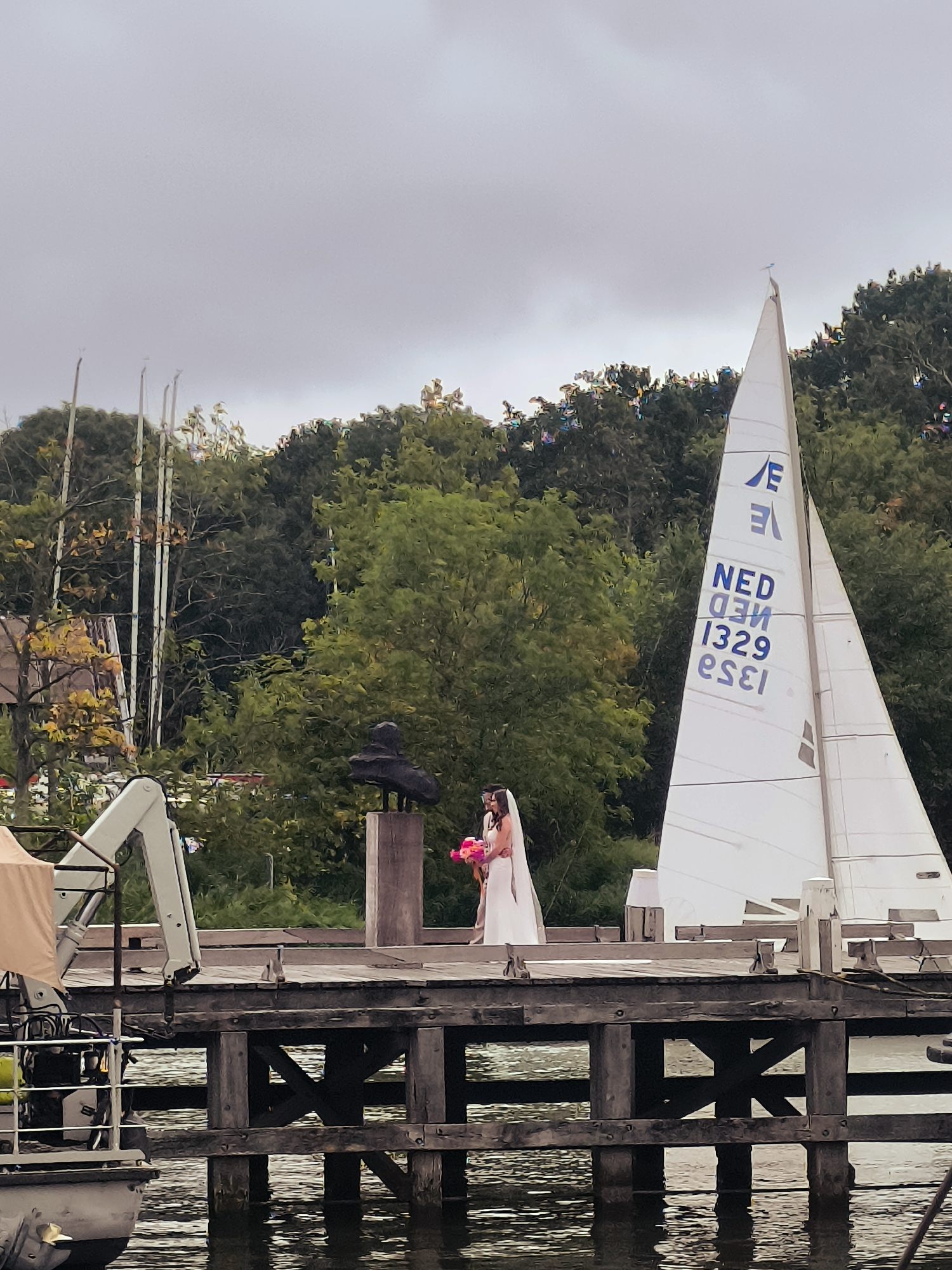 A newlywed couple, a bride in a white wedding dress with a veil and a groom in a suit, stands on a wooden pier in Hoorn, Netherlands. The bride is holding a pink bouquet. A white sailboat with the markings "NED 1329 ESEI" is docked behind them. In the background, there are trees and a cloudy sky. Part of a boat with a crane is visible on the left side of the image. A dark sculpture is mounted on a pedestal on the pier.