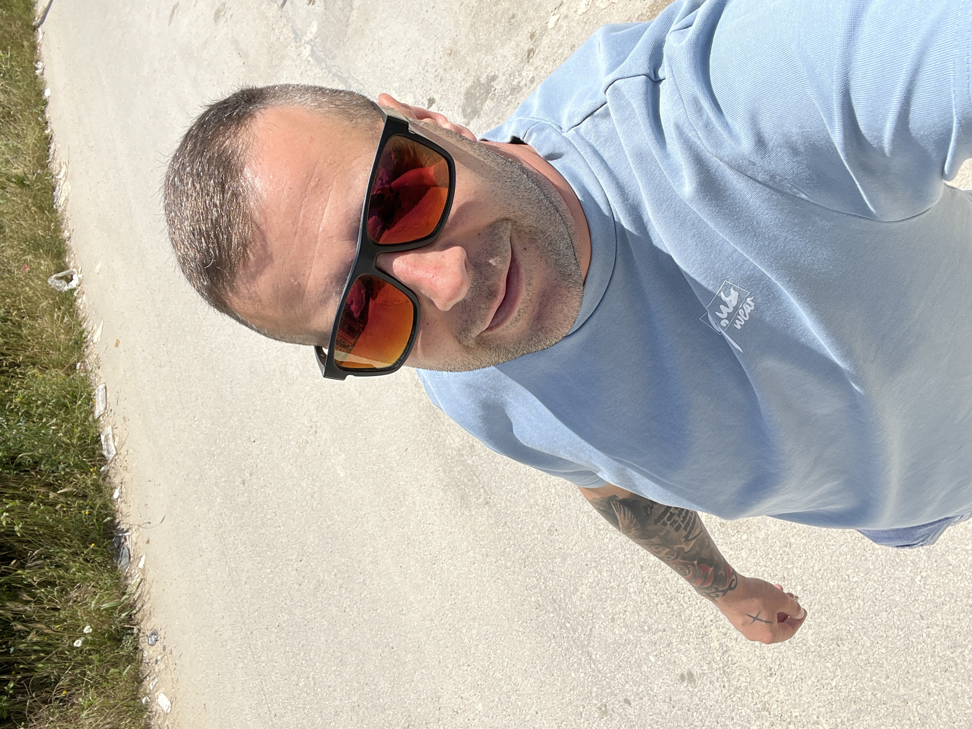 A man wearing sunglasses and a light blue t-shirt is taking a selfie outdoors. He has short hair and stubble. A tattoo is visible on his left arm, and a small cross tattoo is on his left hand. The background is a dirt road with some grass and vegetation. The image appears to be taken in Varna, Bulgaria, as indicated by the presence of a white building with Cyrillic lettering visible in the background.