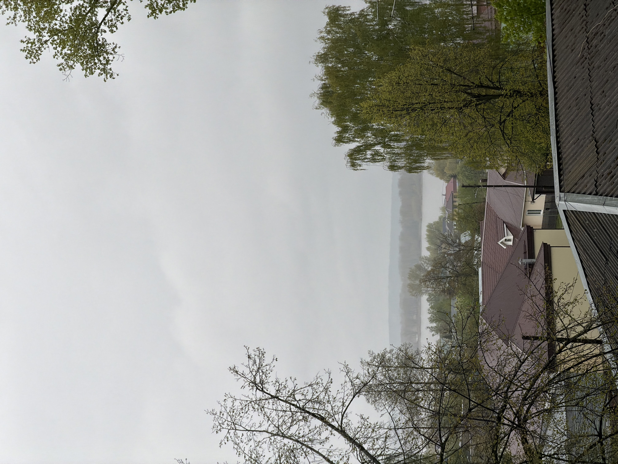 View from above in a rainy, misty area of Chaikovsky, Russia. The scene includes a glimpse of buildings, trees, and a foggy body of water. The foreground contains trees with bare branches, as well as a view of rooftops and parts of buildings. The water, reflecting the sky and trees, is obscured by the fog.