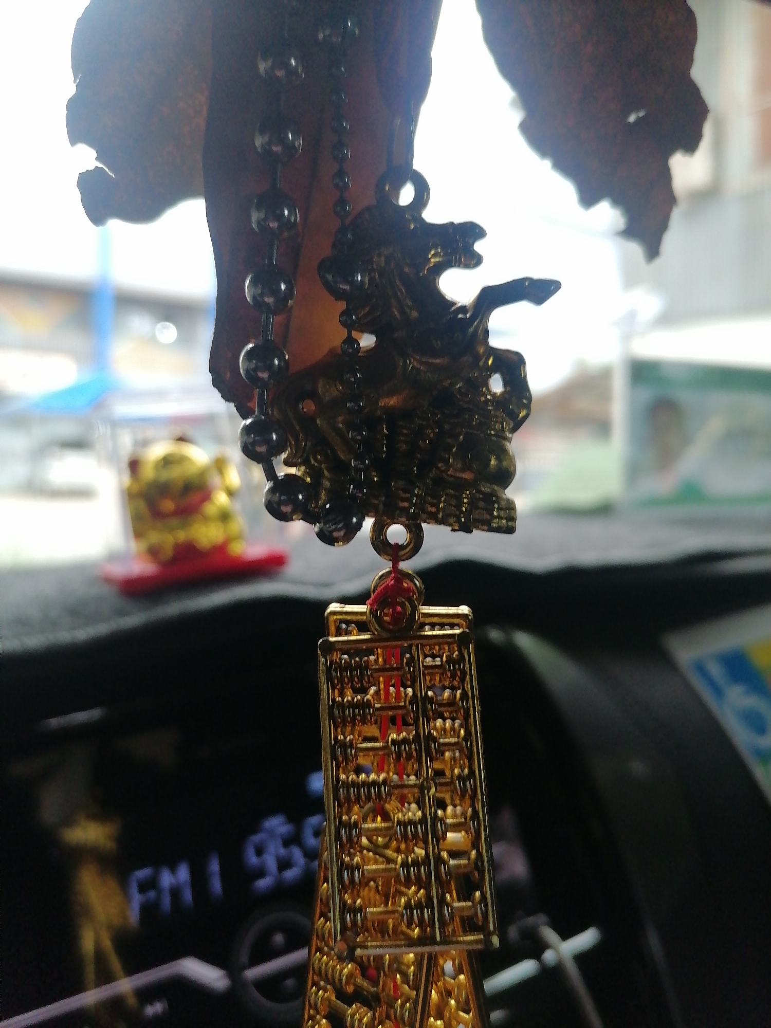 The image depicts a car interior with a hanging ornament. The ornament consists of a beaded chain, a decorative metal dragon, and a rectangular gold-colored piece resembling an abacus. In the background, through the windshield, there is an out-of-focus cityscape, suggesting an urban location. A golden Maneki Neko figurine sits on the dashboard in the background, near the windshield. On the dashboard in the foreground, “FM1 95.9” is visible on a display screen. Given the presence of the Maneki Neko and the “FM1 95.9” radio station, this image was captured in the city of Davao City, Philippines.