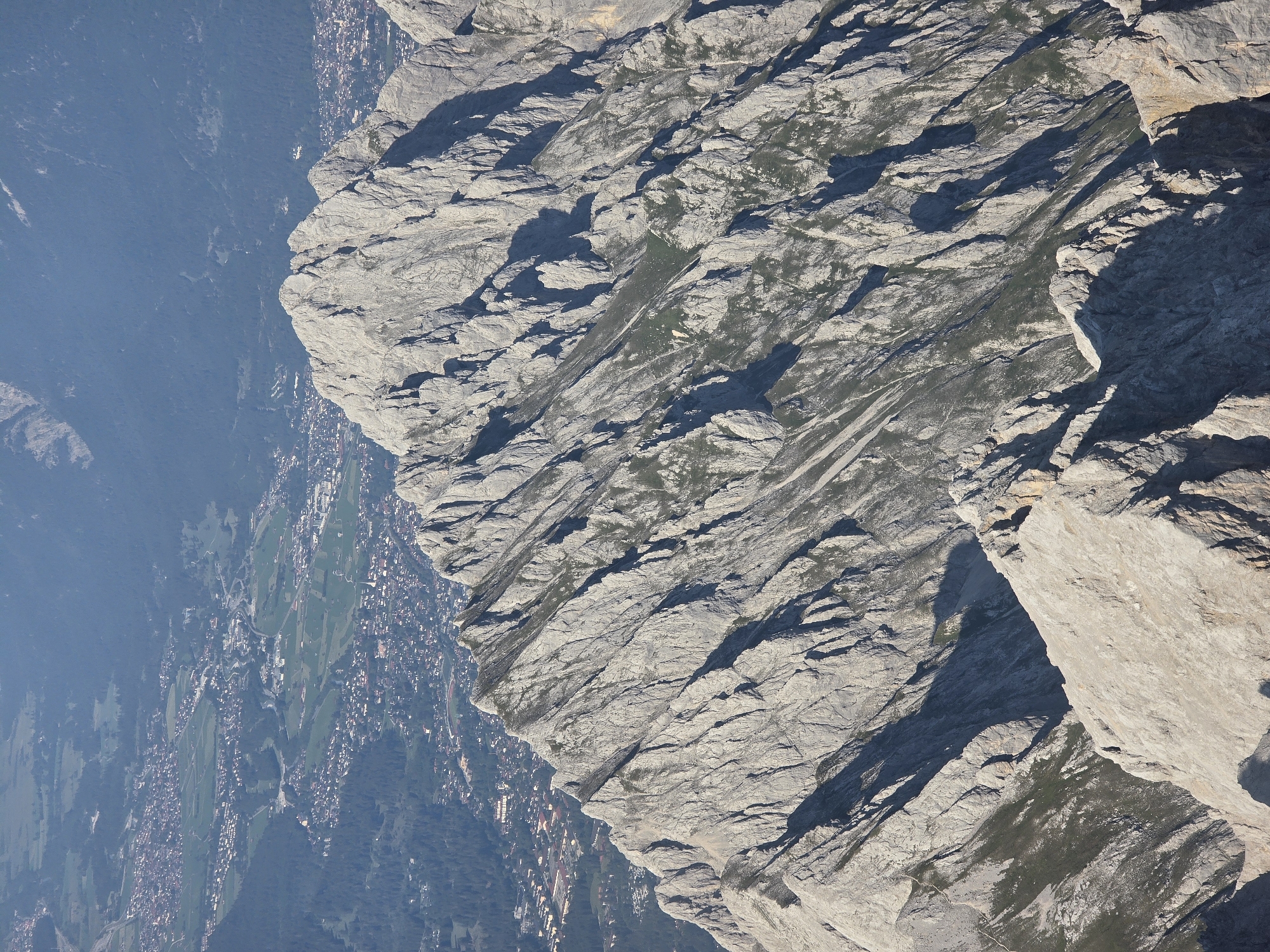 This image is a high-angle, telephoto shot of a rugged mountain range with a town nestled in the valley below. The foreground is dominated by sharp, grey, rocky peaks characterized by steep slopes, deep crevices, and sharp ridges. Patches of sparse green vegetation cling to some of the less steep inclines.
In the midground, the mountain slopes descend into a valley. A town, identifiable by its dense collection of buildings with red-tiled roofs, spreads across the valley floor and up the lower hillsides. The urban sprawl is interspersed with green fields and darker patches of forest. A road or highway is visible snaking through the town.
The background consists of further mountain ranges, appearing softer and more distant, rendered in shades of blue and grey due to atmospheric perspective. The overall impression is one of a dramatic natural landscape juxtaposed with human settlement. The scene appears to be set in the Bavarian Alps, near Grainau, Germany, given the typical architecture and mountainous terrain. There are no people or animals visible in the image. The lighting suggests a sunny day with strong shadows defining the mountain features.
