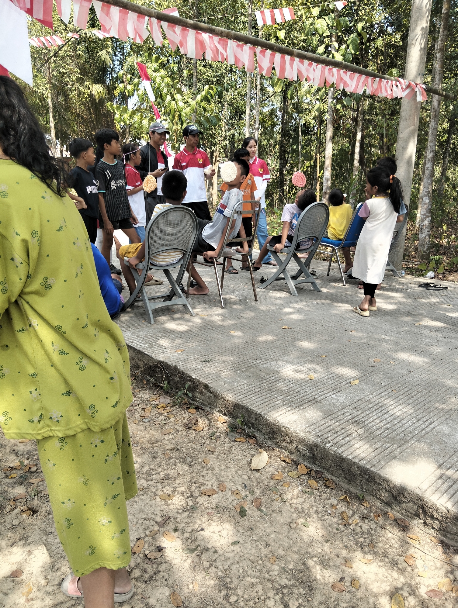 In Sempulang, Indonesia, a group of children and adults are gathered outdoors for a celebration. Red and white Indonesian flags are strung across a bamboo pole above them, decorating the scene. A group of children are seated in chairs, facing away from the camera. To the left, an adult in a bright green patterned outfit stands with their back to the viewer. Other adults and children are standing near a makeshift display with more flags and food items. The ground is a mix of concrete and dirt, with fallen leaves scattered around. The overall atmosphere suggests a community event or a local celebration.