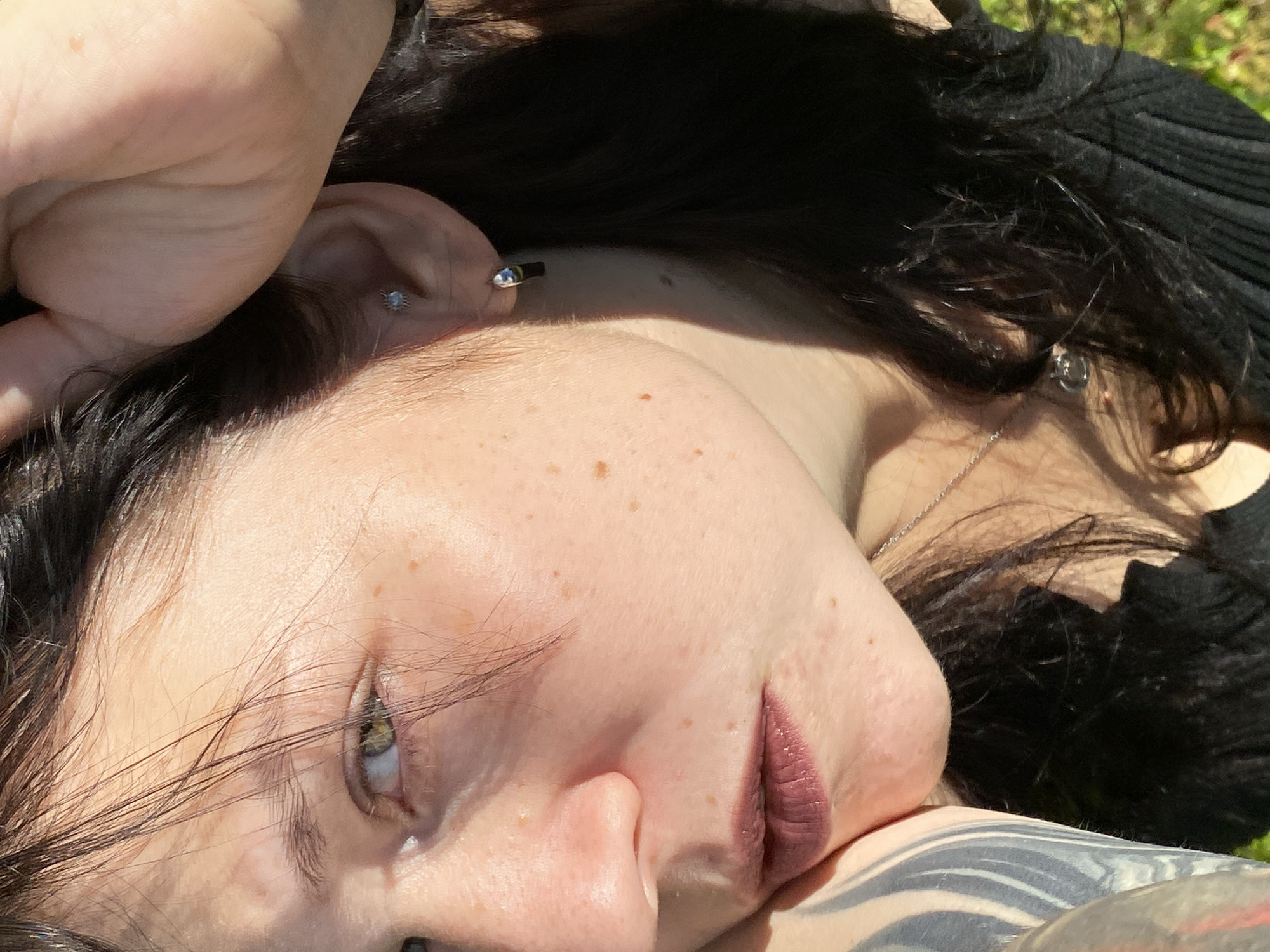 A close-up shot shows a woman's face, turned to the left, with her right hand raised to her head. Her dark hair frames her face, with some strands falling across her forehead and eye. She has light skin with freckles, green eyes, and is wearing dark lipstick. A small earring is visible in her right ear. The background is out of focus, suggesting an outdoor setting with dappled sunlight. There is no visible indication of the city of Фоки, Russia, or any other specific urban environment. The image focuses solely on the woman's features and a portion of her upper body.