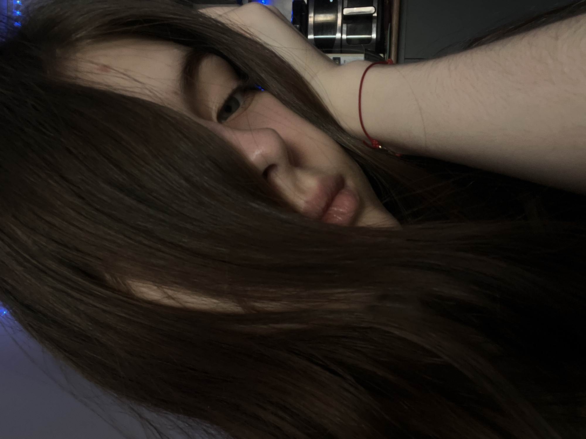 A close-up indoor portrait taken in Saint Petersburg, Russia, depicts an individual with long, dark brown hair falling across the face. The hair covers the left side of the face, partially obscuring the nose and mouth. The visible right eye is light in color. A small red mark is present on the forehead. The person's left hand supports their head, with a red string bracelet visible on the wrist. In the background to the right, metallic kitchen appliances rest on a dark surface, with faint blue ambient light visible in the upper right. The scene is dimly lit.