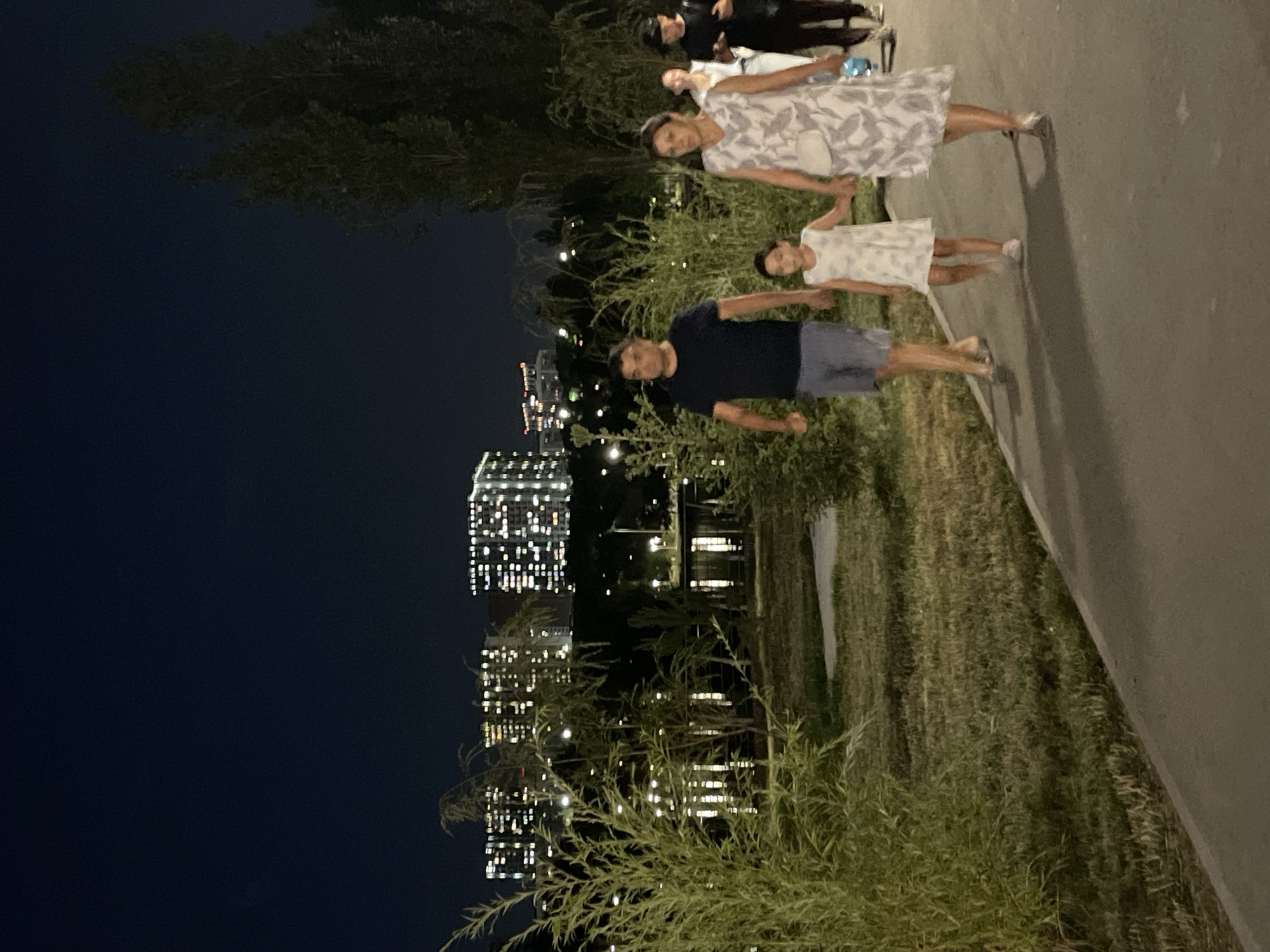 The image depicts a family walking along a paved path in a park at night. In the background, several tall apartment buildings are illuminated with numerous lights, suggesting an urban setting. A body of water, likely a lake or pond, is visible between the park and the buildings, reflecting the lights from the structures. Dense foliage and trees are present throughout the park. A man, a woman, and a young girl are walking together in the foreground, holding hands. The man is wearing a dark t-shirt and blue shorts, while the woman is wearing a light-colored dress with a floral pattern. The girl is wearing a white dress. Two other individuals, a woman and a man, are walking behind them. The overall scene suggests a leisurely evening stroll in a park located in Almaty, Kazakhstan, given the characteristic architecture of the apartment buildings.