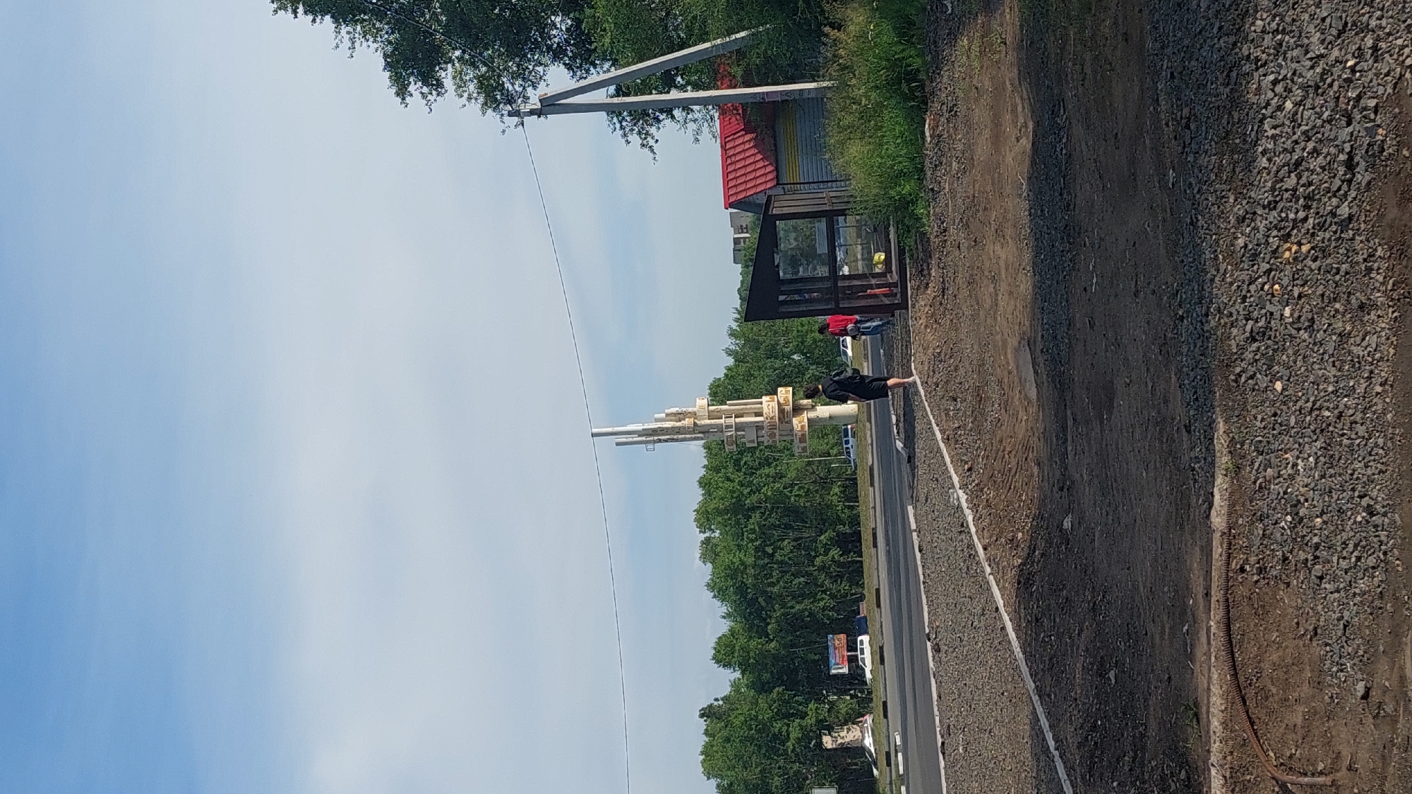 The image shows a bright, clear day with a blue sky and scattered clouds. The scene appears to be an intersection or a roadside area in Amursk, Russia. In the foreground, there is a gravelly, uneven ground with some dirt patches. A rusty, coiled pipe lies on the ground. A white-lined path is visible in the middle ground.
Further back, a road with paved asphalt is present, with some cars driving on it. On the left side of the road, a large, abstract sculpture made of white metallic elements is visible, along with a billboard with Russian text and an image of a white van. On the right side of the road, there is a bus stop with a glass shelter. Behind the bus stop is a small, shed-like structure with a red roof and yellow siding.
Several people are present. A person in a black jacket and shorts stands near the bus stop, facing away from the camera. Two other individuals, one in a red top and the other in a blue top, are standing or walking near the bus stop. A cluster of green trees forms a dense backdrop behind the road and the sculpture. In the distance, a few buildings are visible, suggesting a populated area. The overall impression is that of a public space at the edge of a city.