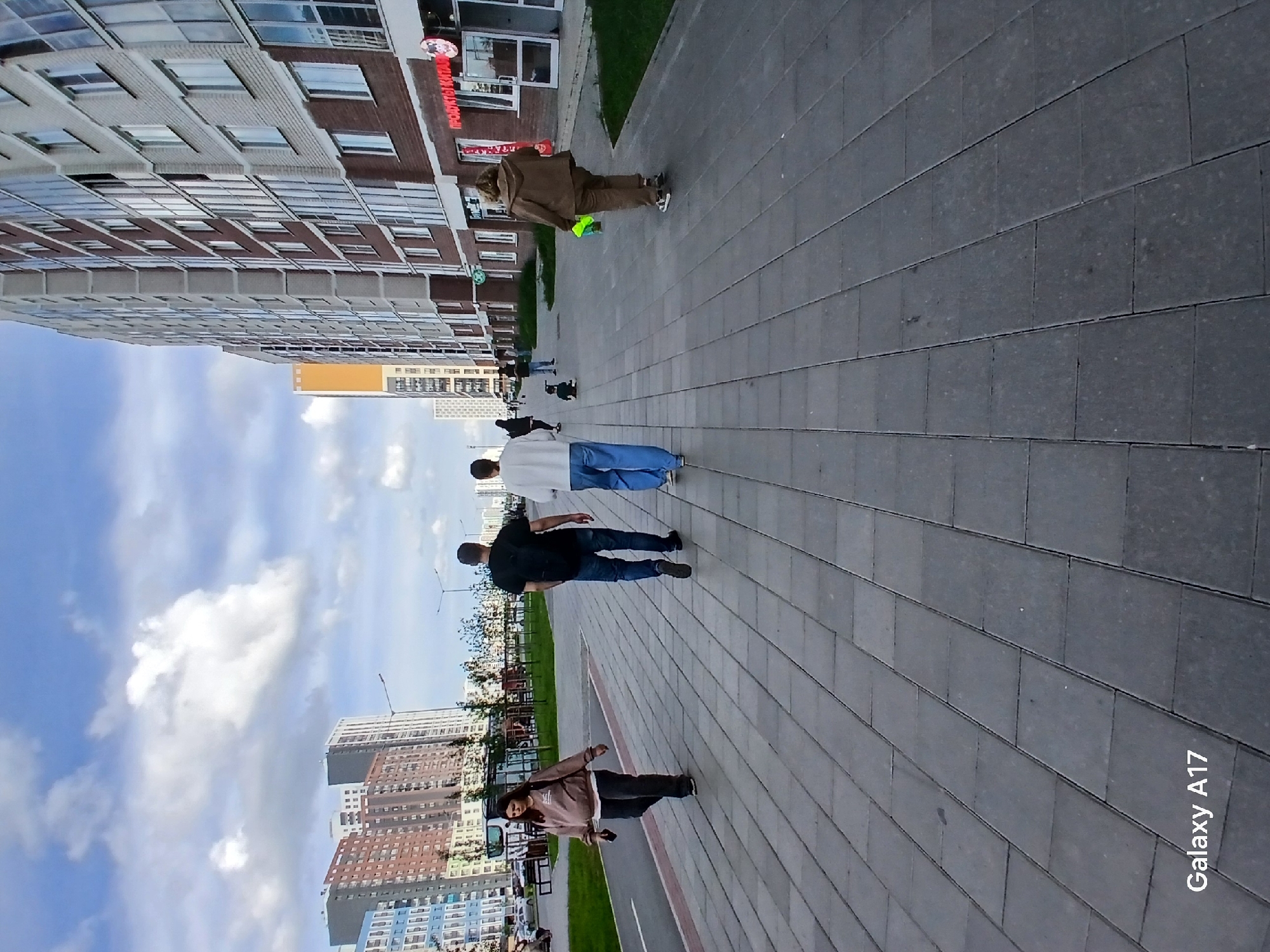 The image shows a paved walkway in an urban environment, possibly Yekaterinburg, Russia. Several people are walking along the path. To the left, there are buildings, including residential structures and what appears to be a retail establishment with signage. The sky is visible with clouds. A "Galaxy A17" watermark is visible in the corner.