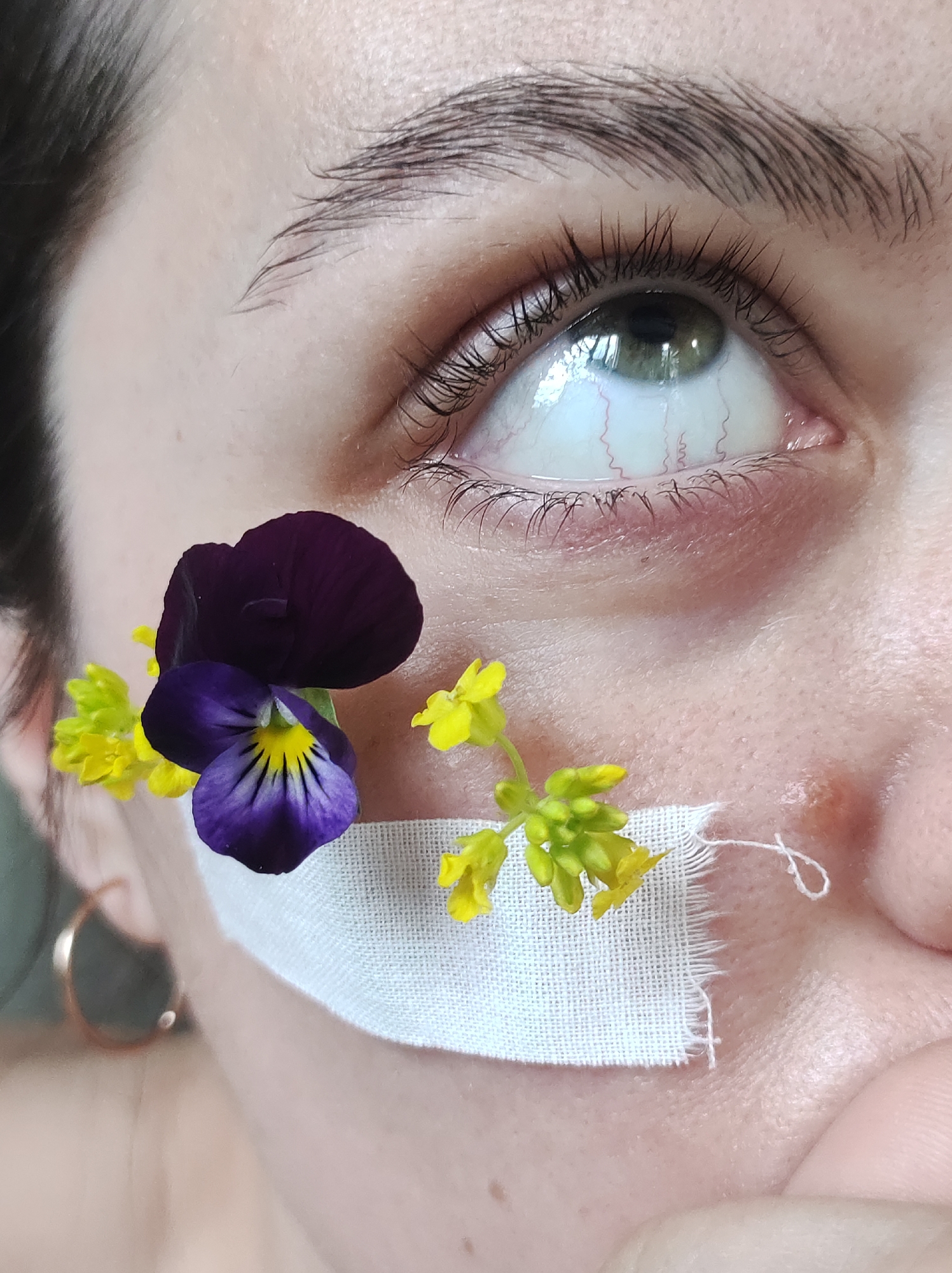 A close-up image focuses on a person's face, specifically the area around their eye and cheek. The eye is light in color and has some visible blood vessels. The eyebrow above the eye is dark and well-defined. A square of white fabric is placed on the cheek, and two small floral arrangements are attached to it. One arrangement features a dark purple and yellow pansy, while the other consists of small yellow flowers. The person's skin is light in tone, and a gold hoop earring is visible in the background. The city is not recognizable in the image.
