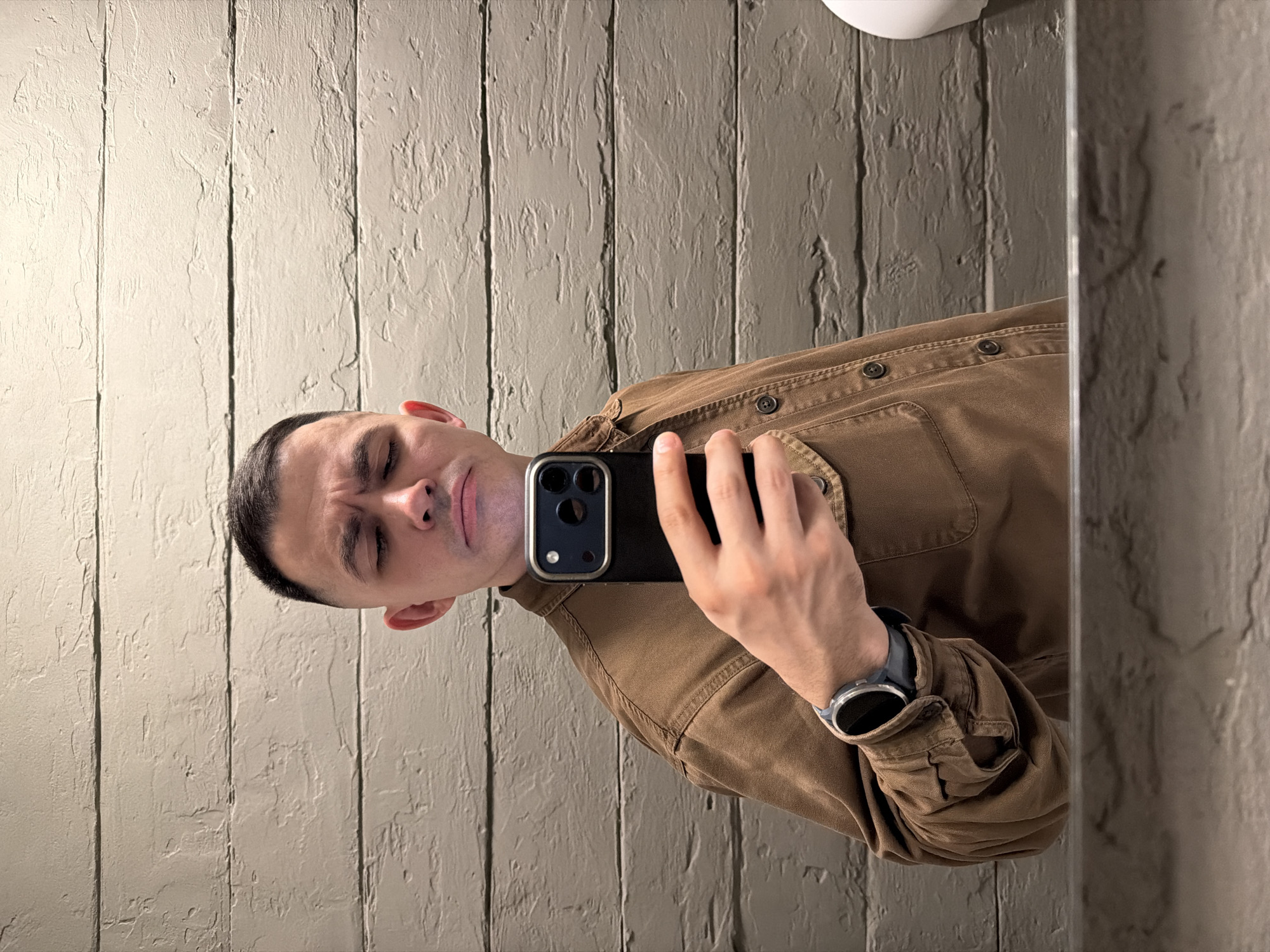 A young to middle-aged man is captured in a mirror selfie, standing in an indoor setting. He has short, dark hair and appears clean-shaven. He is wearing a brown or tan collared long-sleeved shirt with buttons down the front and on the pockets, suggesting a casual yet sturdy material like canvas or denim. A smartwatch with a dark band and silver bezel is visible on his left wrist.
The man holds a black smartphone with a prominent camera module featuring multiple lenses and a gold or bronze colored frame in his right hand, angled towards the mirror to take the photo. His expression is a notable detail, characterized by slightly furrowed eyebrows, narrowed eyes, and a subtle smirk, conveying a somewhat playful, skeptical, or quizzical emotion. His head is tilted slightly to one side.
The background consists of a textured wall, appearing to be composed of vertical panels with a distressed or rough finish, colored in a light brownish-grey tone. The edge of the mirror is visible on the right side of the frame, showing a reflection of the textured wall and part of a white fixture, possibly a light or ceiling element, at the top. The lighting is uniform, suggesting indoor artificial light. There is no visible text in the image.
The photo was taken in Izhevsk, Russia.