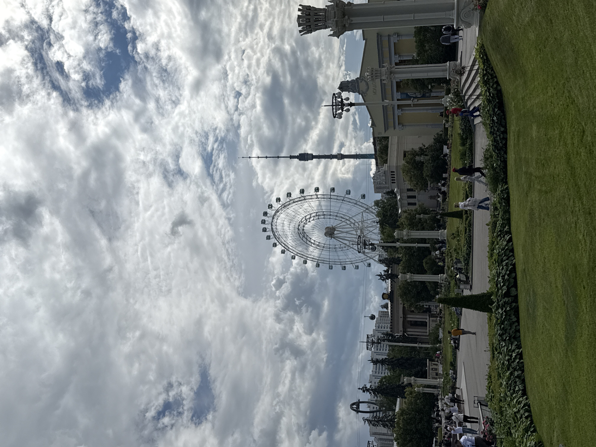 The image depicts an outdoor scene at the Exhibition of Achievements of National Economy (VDNKh) in Moscow, Russia, under a cloudy sky. In the background, a large white Ferris wheel is visible, and to its right, a tall, slender television tower stands out. The foreground features a manicured green lawn and garden beds with lush foliage. Several people are seen walking on pathways and sitting on benches throughout the park. Architectural elements, including classical columns and buildings, are also present. The overall atmosphere suggests a public park or exhibition space, with visitors enjoying the day despite the overcast weather.