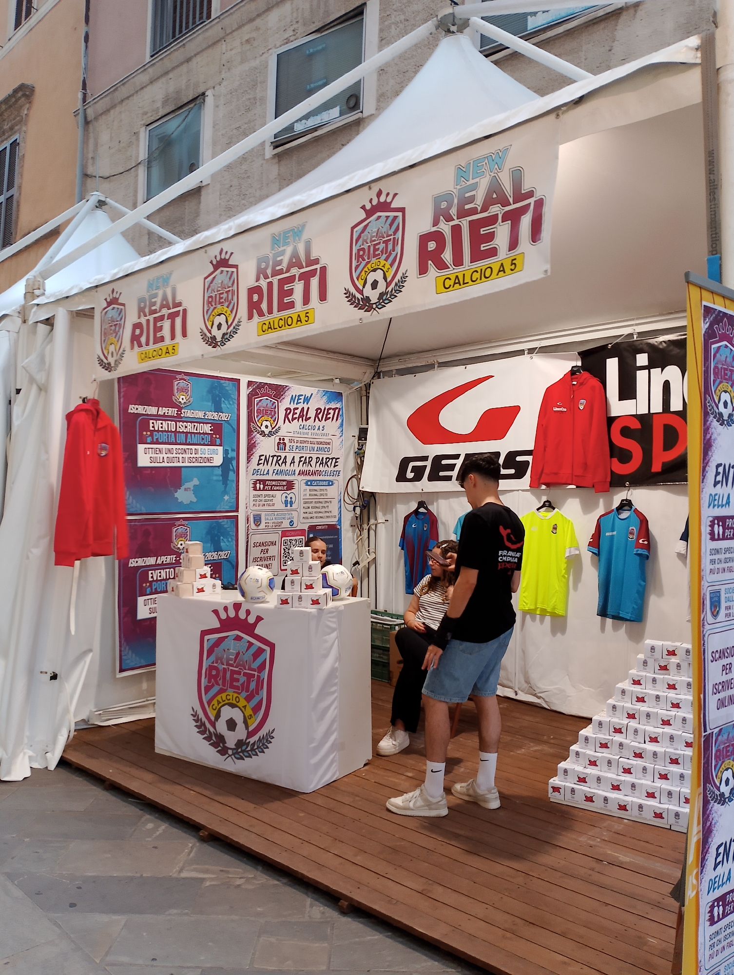 The image depicts an outdoor event stall, likely a promotional booth for "New Real Rieti Calcio A 5," a futsal team based in Rieti, Italy. The stall is set up with white tents and features banners displaying the team's logo and colors, predominantly red and light blue.
Several individuals are present. In the foreground, a young man wearing a black t-shirt, denim shorts, white socks, and sneakers stands facing away from the camera, seemingly interacting with the stall. To his left, a young woman with dark hair, wearing a striped top, sits on a chair, also facing the stall. Further to the left, a red hoodie is displayed on a hanger, potentially for sale or promotion.
The stall itself is adorned with posters and promotional materials. One poster on the left advertises "Iscrizioni Aperte - Stagione 2026/2027" (Open Registrations - Season 2026/2027) and "Evento Iscrizione - Porta un Amico!" (Registration Event - Bring a Friend!). Another poster on the right reiterates "New Real Rieti Calcio A 5" and promotes registration with various offers.
A table in front of the stall is covered with a white cloth featuring the team's logo. On this table, there are several white footballs and numerous small white boxes stacked in a pyramid formation, some of which have the team's logo printed on them.
In the background, clothing items such as t-shirts in various colors (blue, yellow, red) are displayed on hangers. A large banner with a red "G" logo and "GEBS" text is visible on the back wall. The environment appears to be a public space, possibly a town square or pedestrian area, with a building facade visible behind the tents and a wooden platform flooring laid out for the stall. The overall scene suggests a promotional event aimed at recruiting new players or promoting the team.