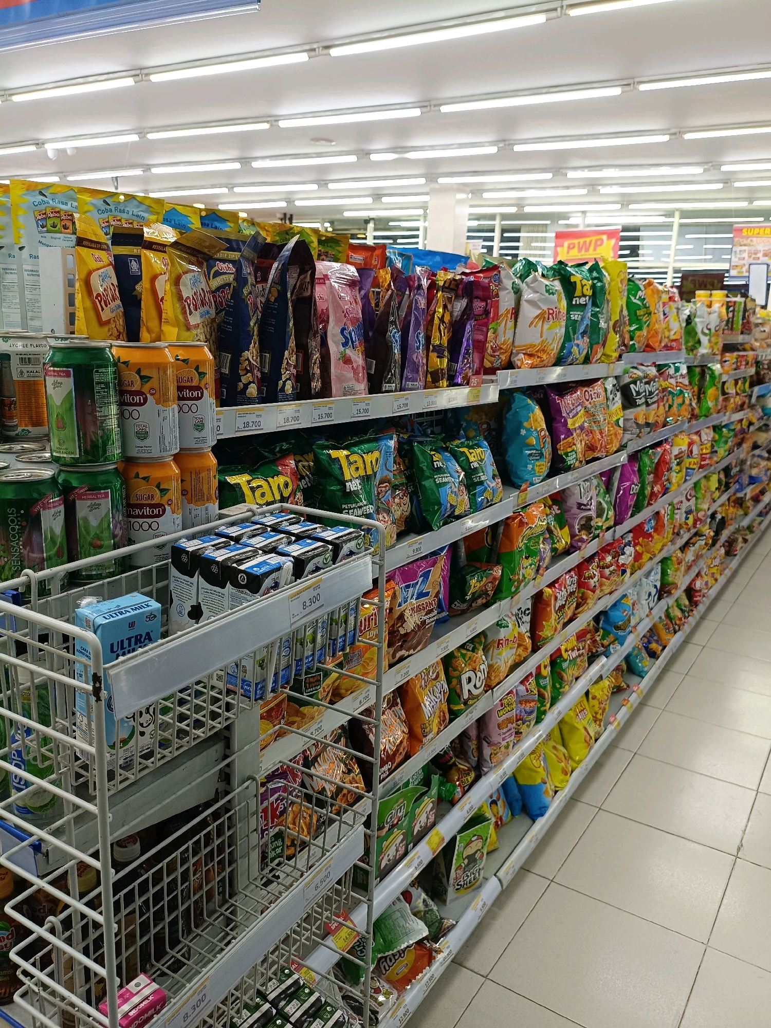 This is a medium shot, eye-level, interior photograph of a convenience store aisle in Maguwoharjo, Indonesia, featuring a wide array of packaged snacks and beverages displayed on white metal shelving units. The shelving is organized into multiple tiers, with products ranging from colorful chip bags and cereal boxes to juice cartons and canned drinks. The products are densely packed, with some items stacked vertically and others arranged horizontally. Prominent branding and text are visible on many of the packages. In the foreground, a white wire rack holds several cartons of "ULTRA MILK" and other unidentified beverages. The lighting is bright and overhead, consistent with a commercial retail environment, and the floor is tiled in a light neutral color. The overall scene depicts a well-stocked retail space with a focus on snack food and drinks.