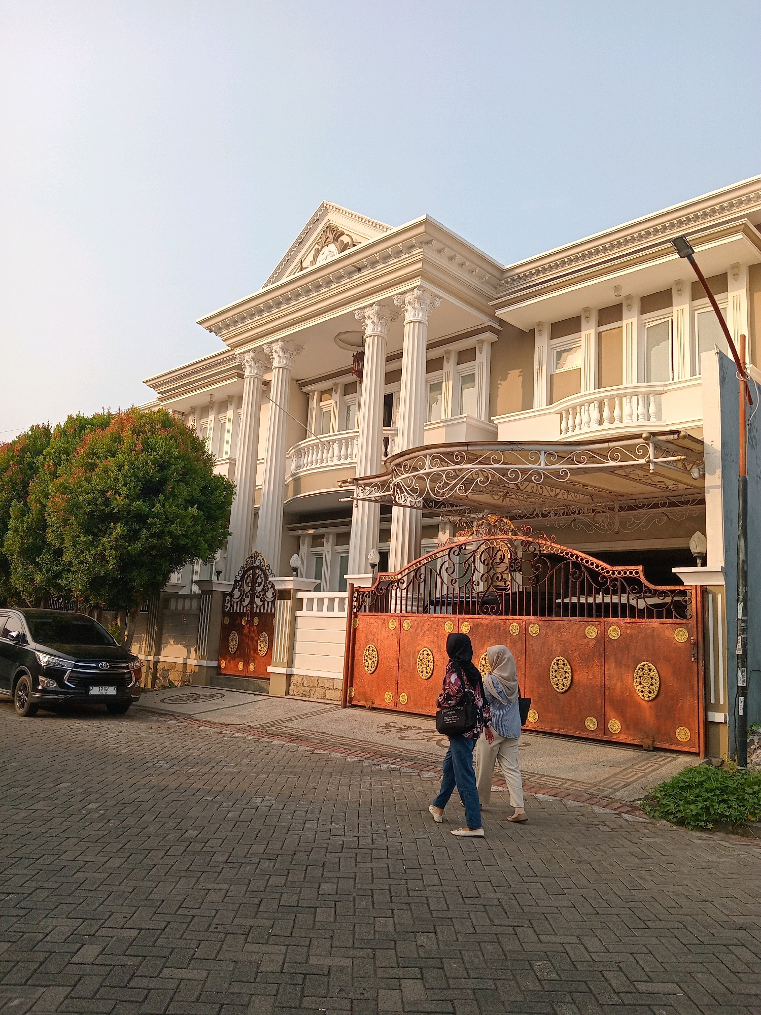 In the city of Randuagung, Indonesia, a stately two-story house with classical architectural elements stands under a clear sky. The house features a prominent portico with Corinthian columns, a decorative frieze, and a balcony with an ornate balustrade. A large, ornate gate, primarily copper-colored with intricate gold embellishments, guards the driveway. To the left of the house, a lush, full green tree with reddish new growth is visible. Parked on the brick-paved driveway is a dark-colored SUV. Two women, both wearing hijabs, are walking on the brick sidewalk in front of the gate. The woman on the left wears a patterned top and blue jeans, carrying a dark bag. The woman on the right is dressed in a light blue striped shirt and light-colored pants, also carrying a dark bag. The environment is residential, with paved walkways and a sense of well-maintained property.