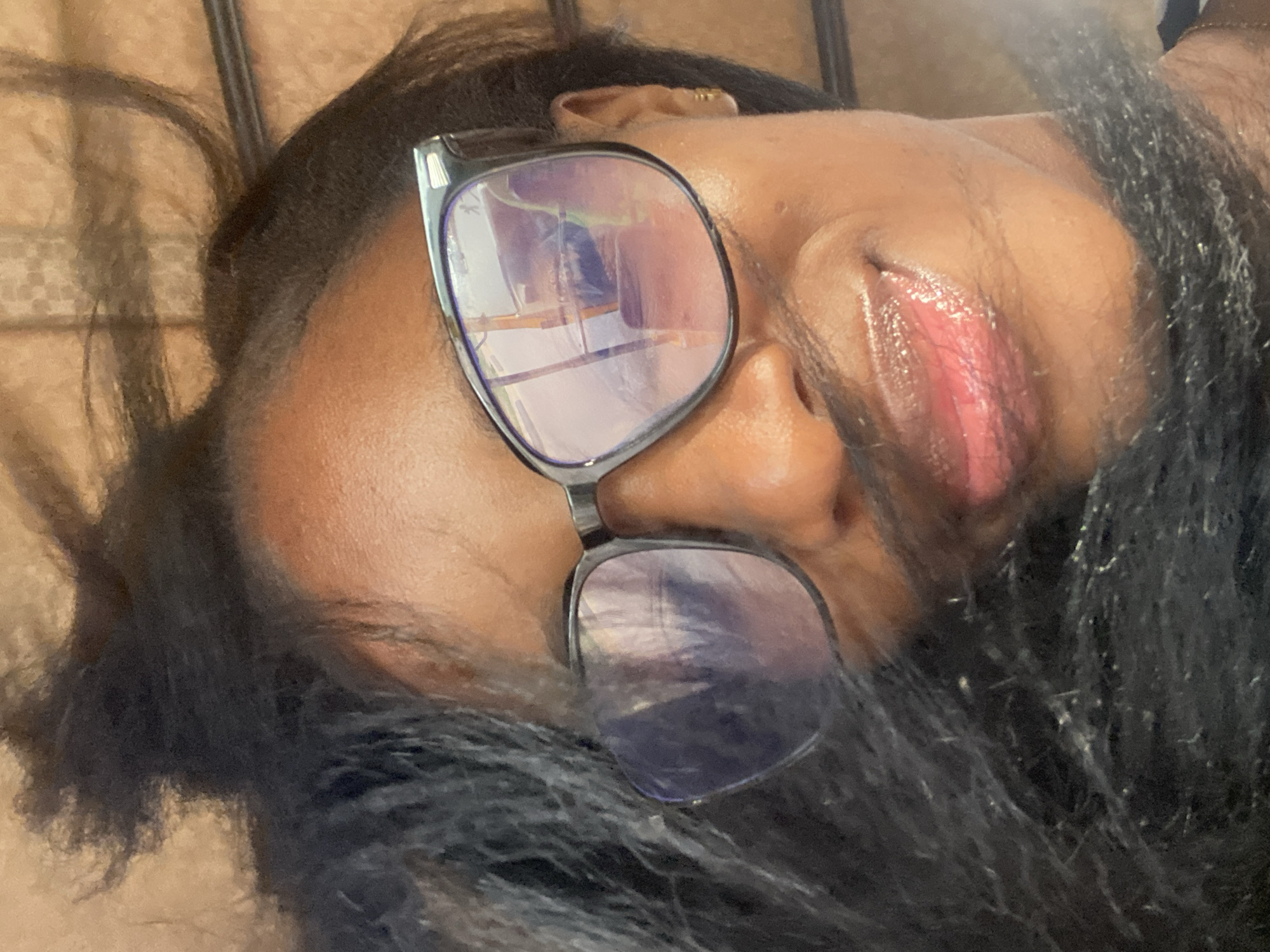 The image is a close-up, over-the-shoulder selfie taken by a Black woman in Gura Top, Nigeria. She is wearing large, dark-rimmed glasses and has dark, long hair that frames her face. Her lips are glossed and slightly parted, and she appears to be smiling. The background is blurred and suggests an indoor setting with textured walls. The lighting is warm and highlights her skin.