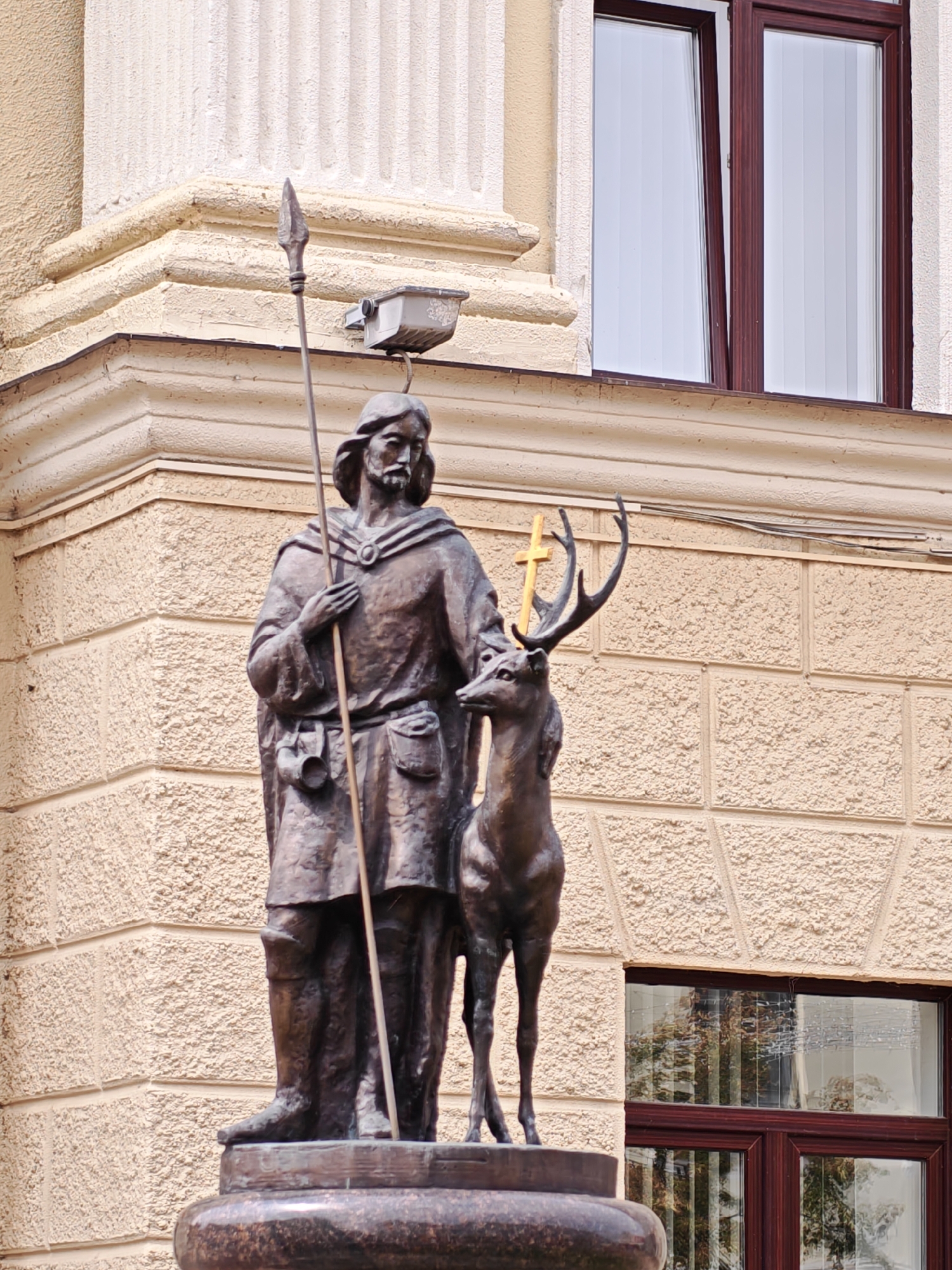 The image shows a bronze statue of a man with a spear and a deer, situated in front of a building with a textured facade. The man has long hair and a beard, and is wearing a robe and boots. He is holding a spear in his right hand. The deer is standing next to him, with a golden cross on its antlers. The building has a large window with blinds on the right side and architectural details above the statue. The statue is located in Hrodna, Belarus, and is believed to be a depiction of Saint Hubertus, the patron saint of hunters.