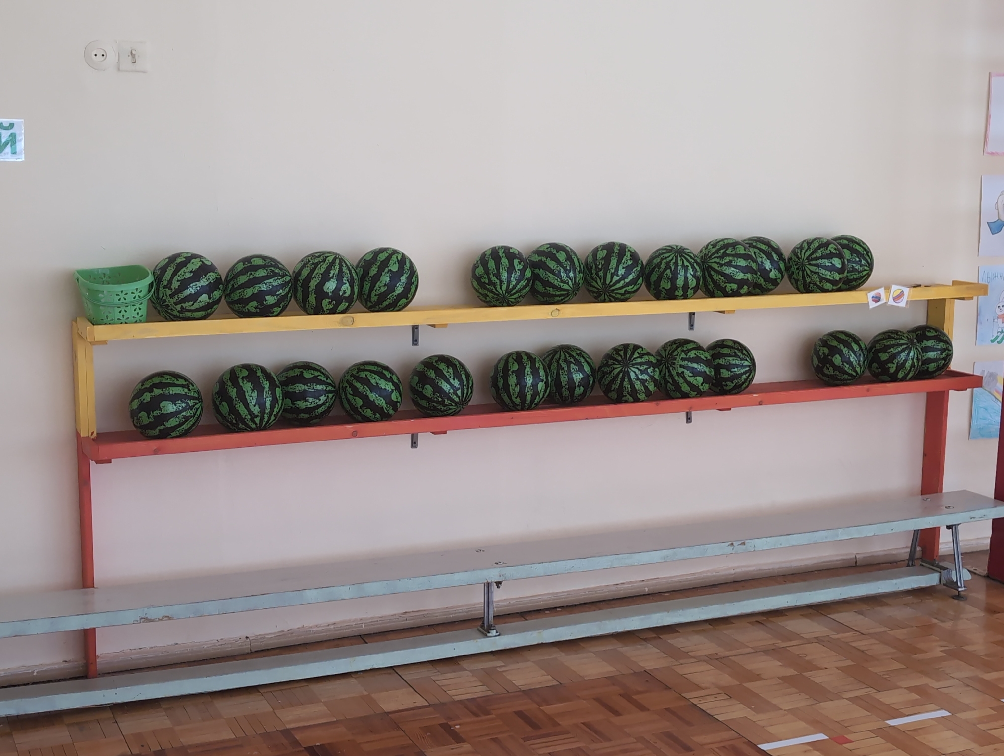 This image captures an indoor scene, likely within a children's facility such as a kindergarten or school in Izhevsk, Russia. The primary subjects are numerous watermelon-patterned balls neatly arranged on a multi-tiered shelving unit, along with a light blue bench and a plain wall.
The shelving unit consists of three levels. The top shelf, painted yellow, holds a small green plastic basket on its far left, followed by twelve black and green watermelon-patterned balls. The middle shelf, painted red, also displays twelve identical watermelon balls. Similarly, the bottom red shelf contains another row of twelve watermelon balls. In total, thirty-six such balls are visible on the shelves.
Below the shelving unit, a long, light blue wooden bench extends across the frame. The floor is made of parquet wood, indicating an indoor setting. The wall in the background is a neutral light color, featuring two electrical outlets on the upper left. A partial green sign with Cyrillic letters, including "Й", is visible to the left of the shelves. On the far right side of the wall, several children's drawings are displayed, one of which clearly shows the Cyrillic word "ЛЫЖНЫЙ" (ski).
The room is well-lit, suggesting it is daytime. No people are present, and no specific activity is taking place; the arrangement appears to be a storage or display of play equipment. The scene conveys a sense of order in a functional space designed for children.