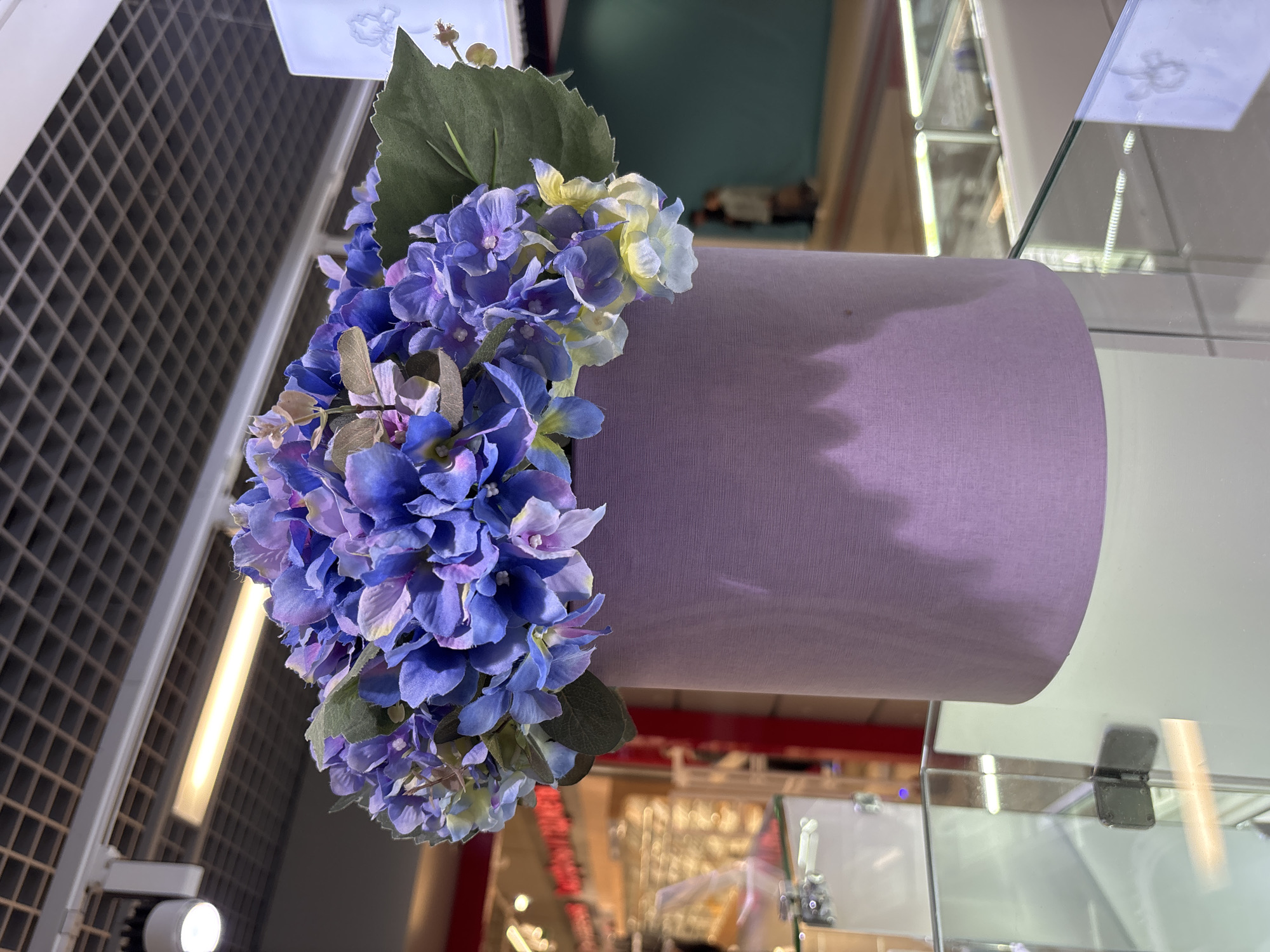 A medium shot focuses on a cylindrical lampshade adorned with purple and green flowers. The lampshade is a light lavender or pale lilac color with a textured appearance resembling fabric. The flowers are predominantly purple, with some lighter yellow-white flowers mixed in. The arrangement is lush and extends out from the side of the lampshade.
The background is a complex array of structures and lights, suggesting an indoor, possibly commercial, environment. A white grid-like structure is visible on the left, indicating a wall or partition. Reflections and glass surfaces indicate display cases or windows. The location is not evident from the image.