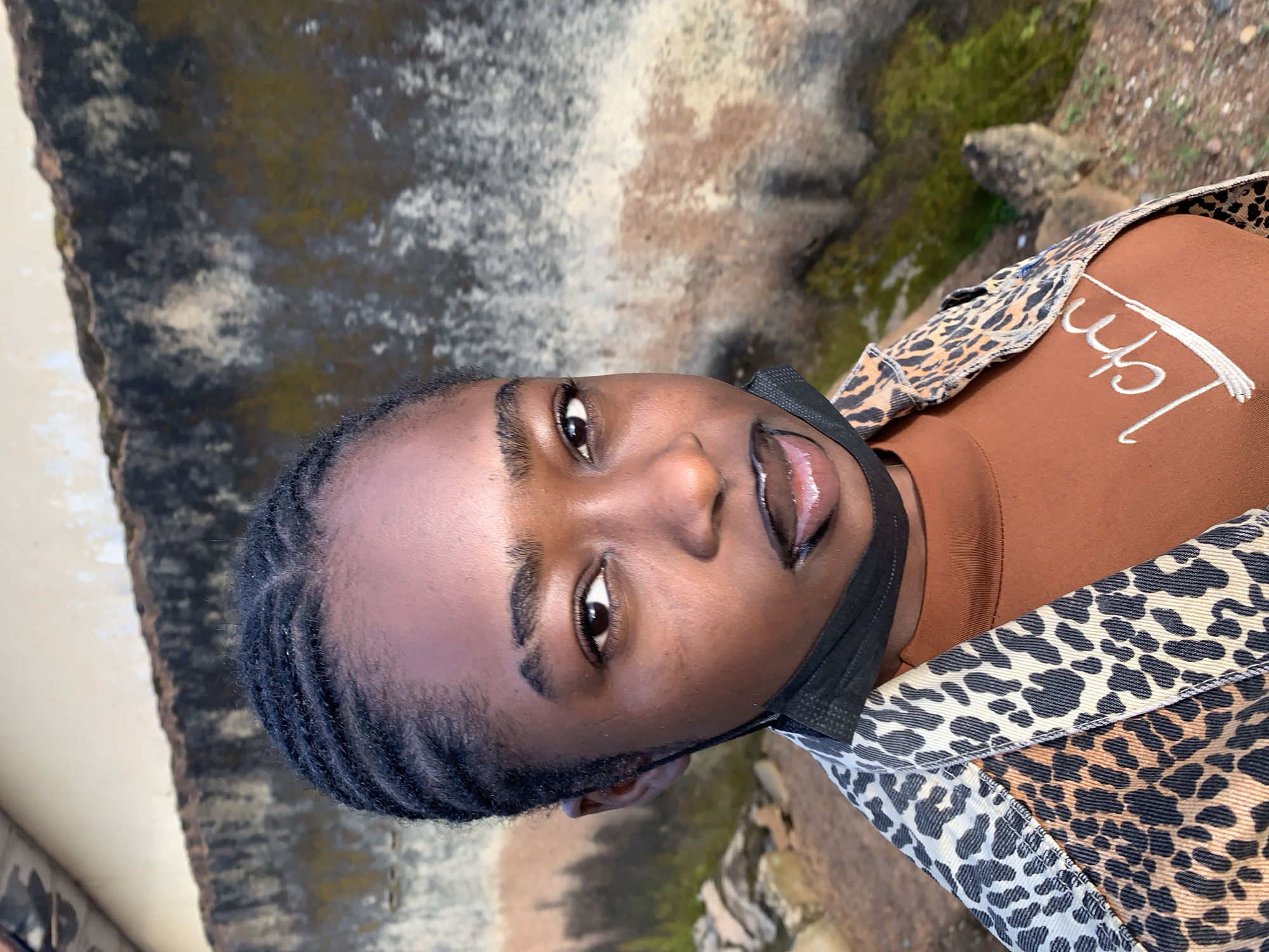 A young Black woman with braided hair and a leopard-print jacket over a brown shirt poses for a photo outdoors. She is wearing a black face mask below her chin and has dark lipstick on. The background consists of a textured concrete wall with some moss and discolored patches. The ground is rocky and dirt-covered. The image appears to be taken during the day. The location is specified as Rafin Par, Nigeria.