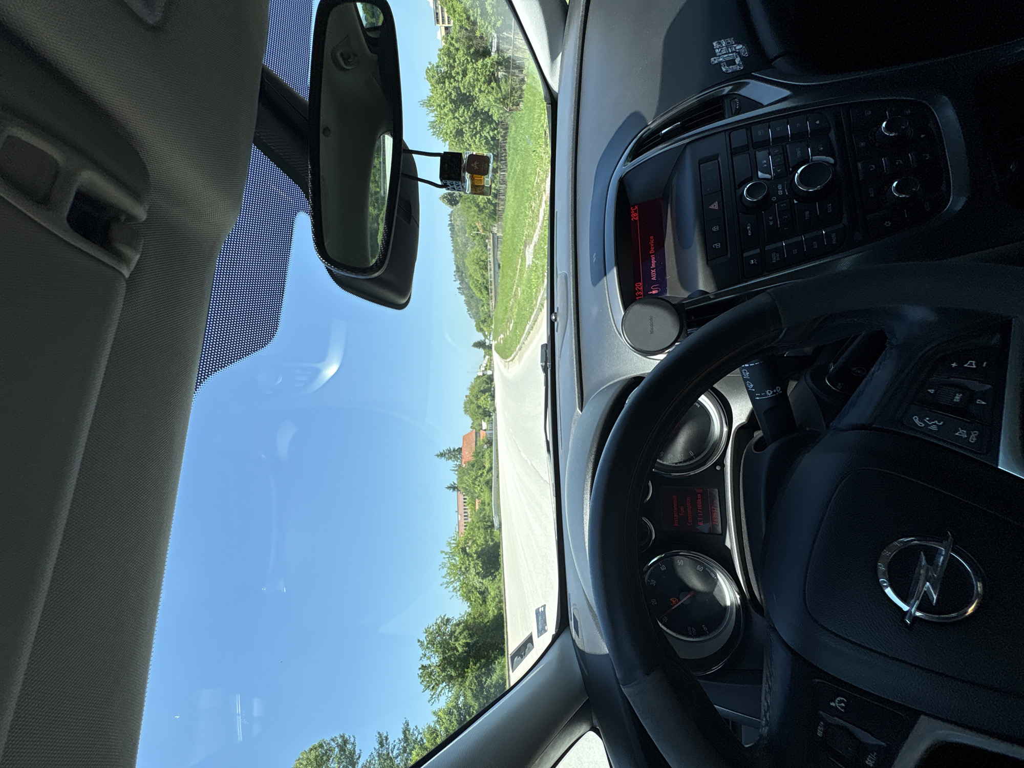 The image shows the interior of an Opel car, specifically the driver's seat. The view is through the windshield, revealing a road leading into a suburban area with houses and greenery, possibly in Ugarchin, Bulgaria, given the context. The dashboard is visible, including the steering wheel with the Opel logo, the instrument panel showing speed and fuel consumption, and the central console displaying temperature and audio information. A phone holder is attached to the dashboard. A rearview mirror is present. The sky is clear and blue.