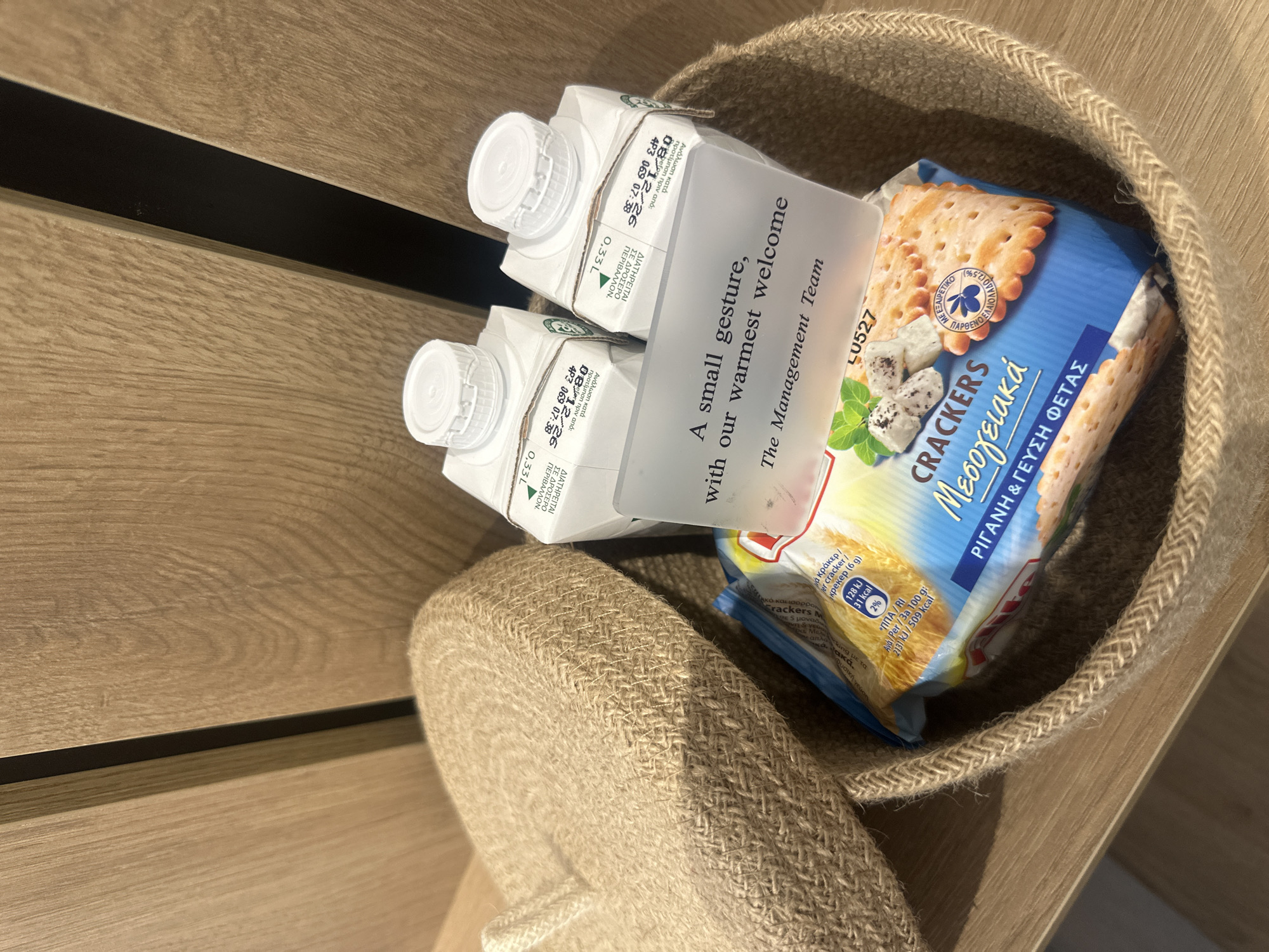 In Larnaka, Cyprus, a woven basket sits on a wooden surface, possibly a shelf or table. The basket contains two small cartons of liquid, likely milk or juice, with white caps and labels printed in Greek. Next to these, a package of crackers is visible, featuring text also in Greek. A transparent plastic card is propped up, displaying a message in English: "A small gesture, with our warmest welcome, The Management Team." The background consists of textured wooden panels.
