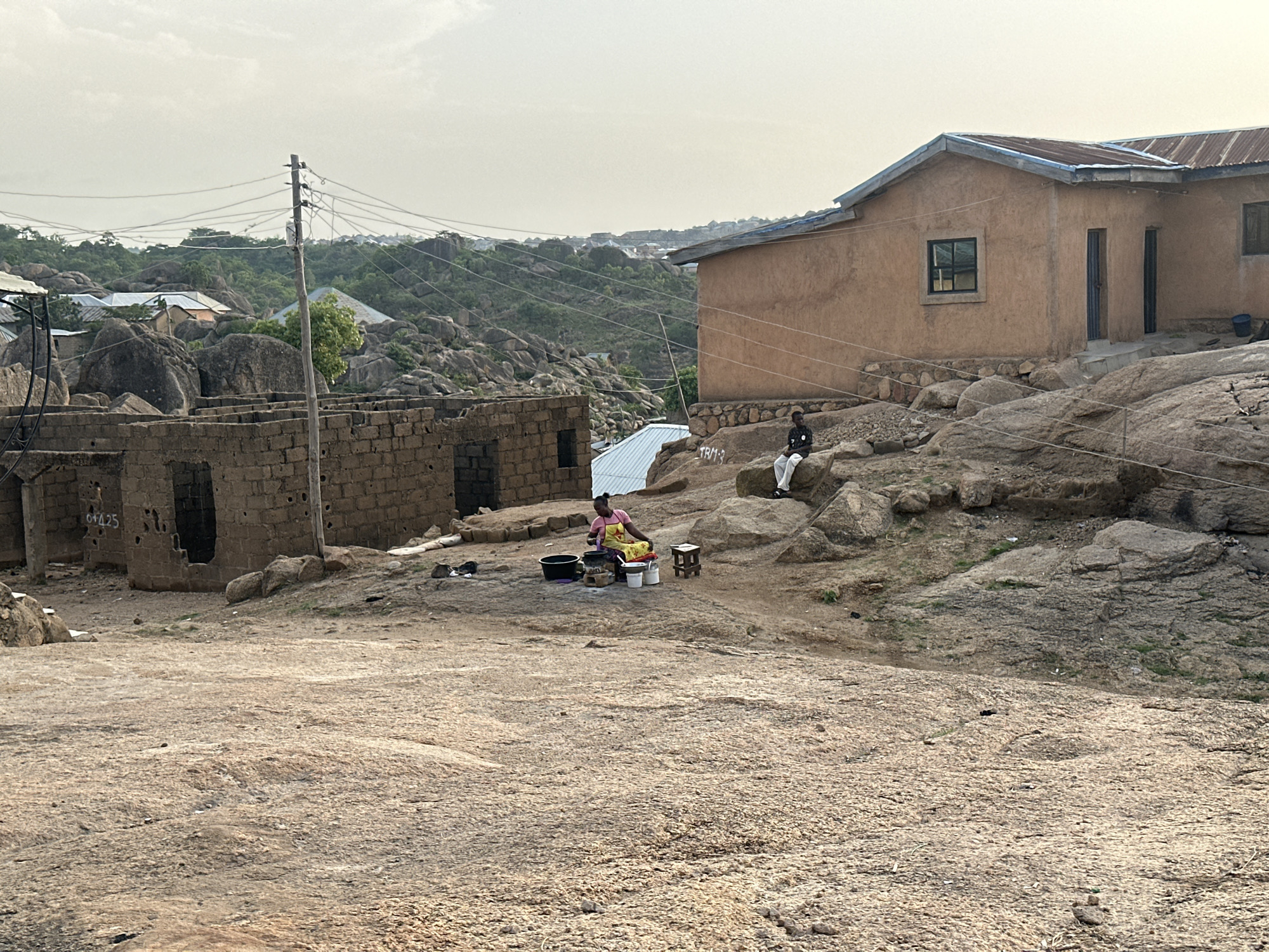 The image shows a scene in Jos, Nigeria, characterized by rocky terrain and sparse vegetation. In the foreground is a large, uneven rock surface that takes up most of the lower half of the frame. Further back, there is a woman wearing a pink shirt and yellow apron, sitting on the ground. She is surrounded by buckets, a pot, and a small wooden stool. To the right of the woman, a man in a dark shirt and light pants is sitting on a rock.
Behind the woman and the man are two partially constructed or dilapidated structures made of dark bricks. In the background, there is a finished building with a metal roof. Power lines run overhead. The overall lighting suggests it is either early morning or late afternoon. The landscape is dotted with large rocks and sparse green vegetation.