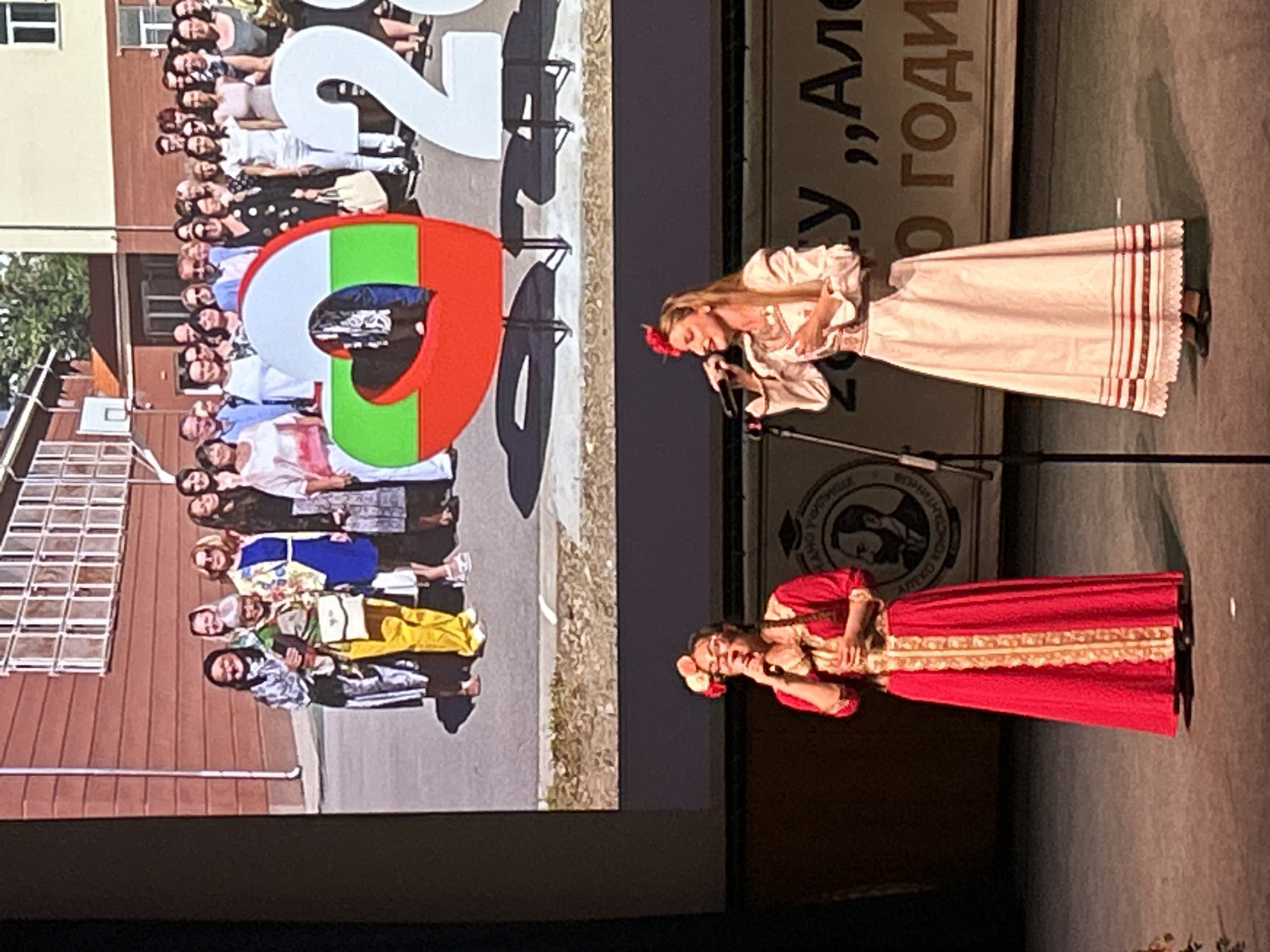 A photograph depicts a stage performance viewed on a large screen. Two women stand at microphones, wearing traditional Bulgarian folk costumes. The woman on the left is dressed in white, and the woman on the right is dressed in red. The screen behind them displays a photo of a group of people posing behind the numbers '2023', with the '2' painted in the Bulgarian flag colors. The group picture is taken outdoors at a school location in Sofia, Bulgaria. The performance appears to be related to a school graduation event.