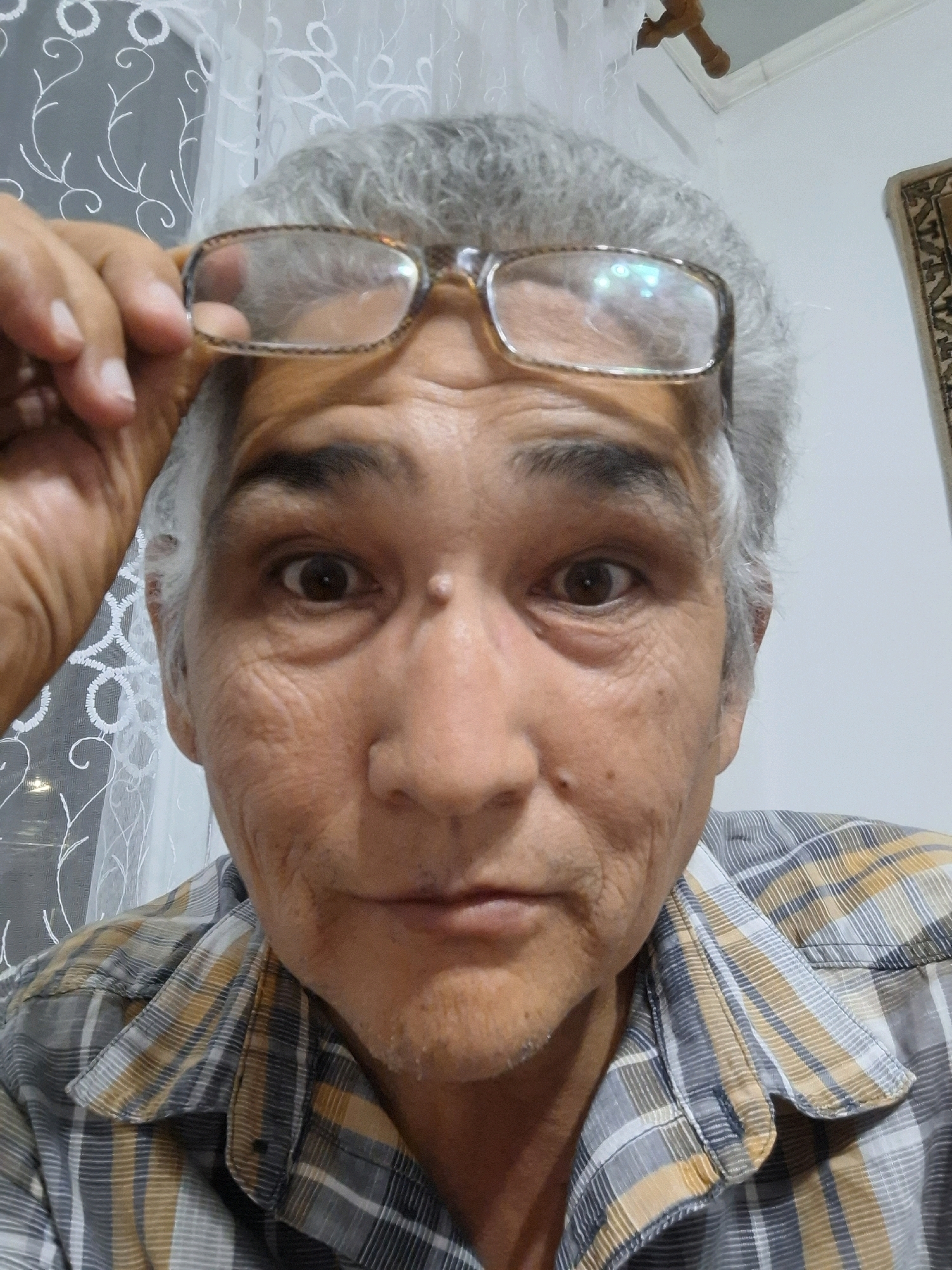 A close-up, low-angle selfie shows an elderly person with gray hair and wrinkles on their face. They are wearing glasses that they are holding up with their left hand. Their eyes are open and looking at the camera. The person is wearing a plaid shirt. The background is out of focus but appears to be indoors with a white curtain or screen with a floral pattern. A small wooden object is visible hanging from the ceiling in the upper right corner. The scene does not contain any specific landmarks or elements that would allow for determination of a location in Almaty, Kazakhstan.