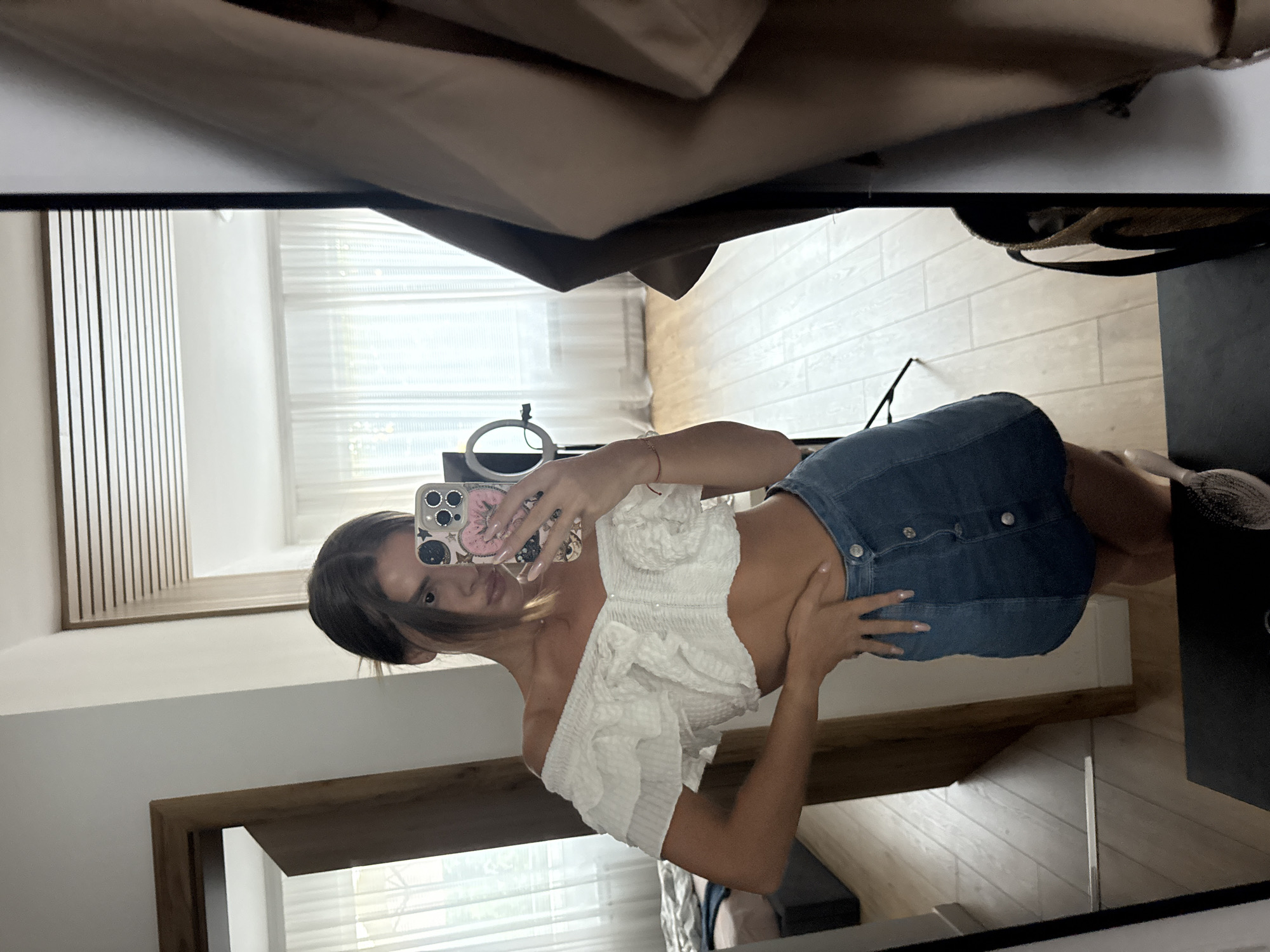 A young woman with dark hair tied back is taking a selfie in a mirror. She is wearing a white, off-the-shoulder ruffled crop top and a denim mini-skirt. Her left hand is on her hip, and her right hand holds her phone, which has a patterned case. Behind her, a room is visible in the mirror's reflection, with a bed, sheer curtains, and wooden floorboards. The setting appears to be an apartment in Plovdiv, Bulgaria.