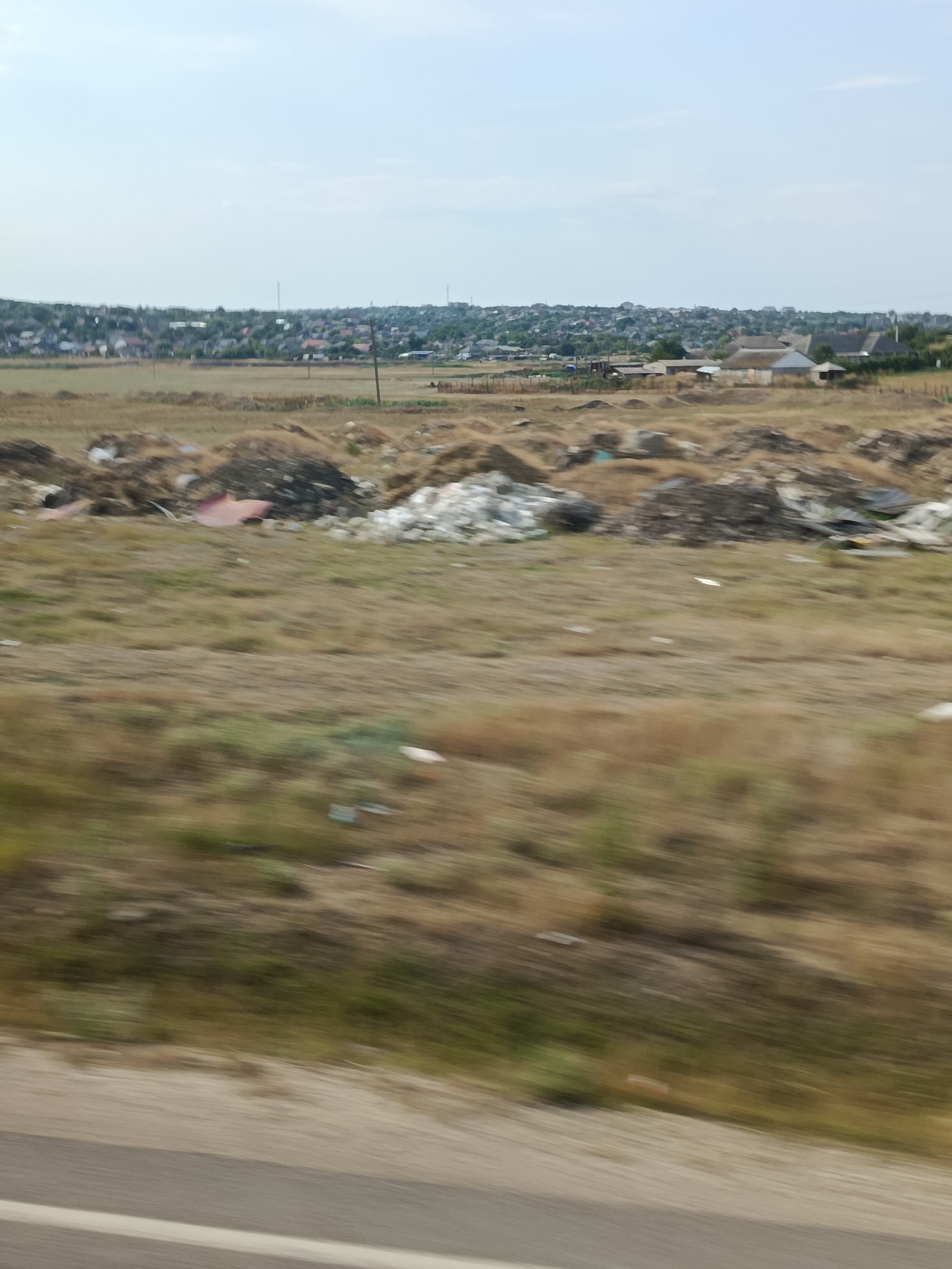This image depicts an outdoor scene with a view of a village or town in the distance, set against a clear, pale blue sky. The foreground is dominated by a dry, grassy field with scattered piles of debris, suggesting a possibly neglected or unmanaged area. The debris includes what appears to be refuse, such as plastic sheeting and other miscellaneous waste, intermingled with dark earth and vegetation.
In the middle ground, a collection of structures, possibly houses or agricultural buildings, are visible. These buildings have varied roof styles, with some appearing to be made of thatch or similar natural materials, while others have more conventional roofing. The village in the background shows a dense collection of buildings with many rooftops, suggesting a populated area. Tall poles, possibly utility poles or radio towers, are also scattered throughout the landscape, both near the village and in the middle ground.
The overall impression is one of a rural or semi-rural landscape under a bright sky, possibly during daytime, given the lighting. The presence of debris in the foreground contrasts with the more organized appearance of the village in the distance. There is no discernible text within the image. The scene appears static, with no specific activity clearly indicated. The motion blur suggests the image was taken from a moving vehicle, likely a road, as indicated by the blurred grey and white stripes at the very bottom of the frame.