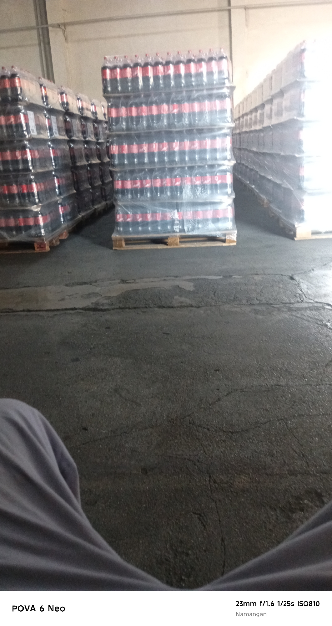 The image depicts a warehouse interior in Namangan City, Uzbekistan. Stacks of bottled beverages, likely Coca-Cola due to the red and black branding on the plastic wrapping, are organized on wooden pallets. These stacks are arranged in rows, with some reaching a significant height. The floor is asphalt, showing signs of wear and cracking. In the foreground, a portion of a person's legs, covered by gray fabric, is visible, indicating the photographer's presence. The lighting suggests an indoor environment, possibly with natural light from a large opening or artificial illumination from overhead. The overall scene conveys a sense of industrial storage and distribution.