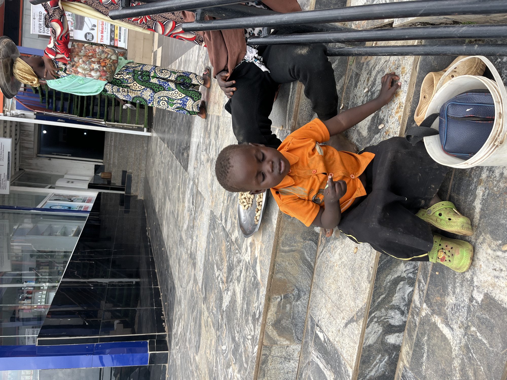 In the city of Galadimawa Village, Nigeria, a young child sits on a set of stone steps, wearing an orange shirt and black pants. The child is holding what appears to be a small snack, possibly nuts, from a metal dish placed on the steps. To the child's right, an adult's legs are visible, suggesting they are sitting next to the child. Nearby, a white bucket contains a blue bag and a pair of light-colored shoes. In the background, other people are seen walking, and a store with reflective windows is visible. The ground is paved with patterned stone tiles.