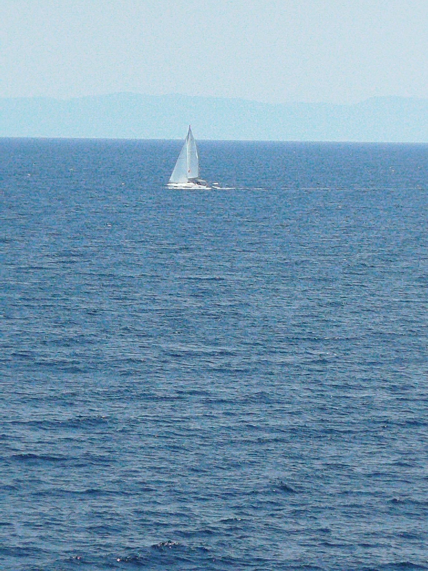 A white sailboat is sailing on the blue sea. In the distance, a hazy outline of mountains is visible against a pale blue sky. The sea is rippled with waves.