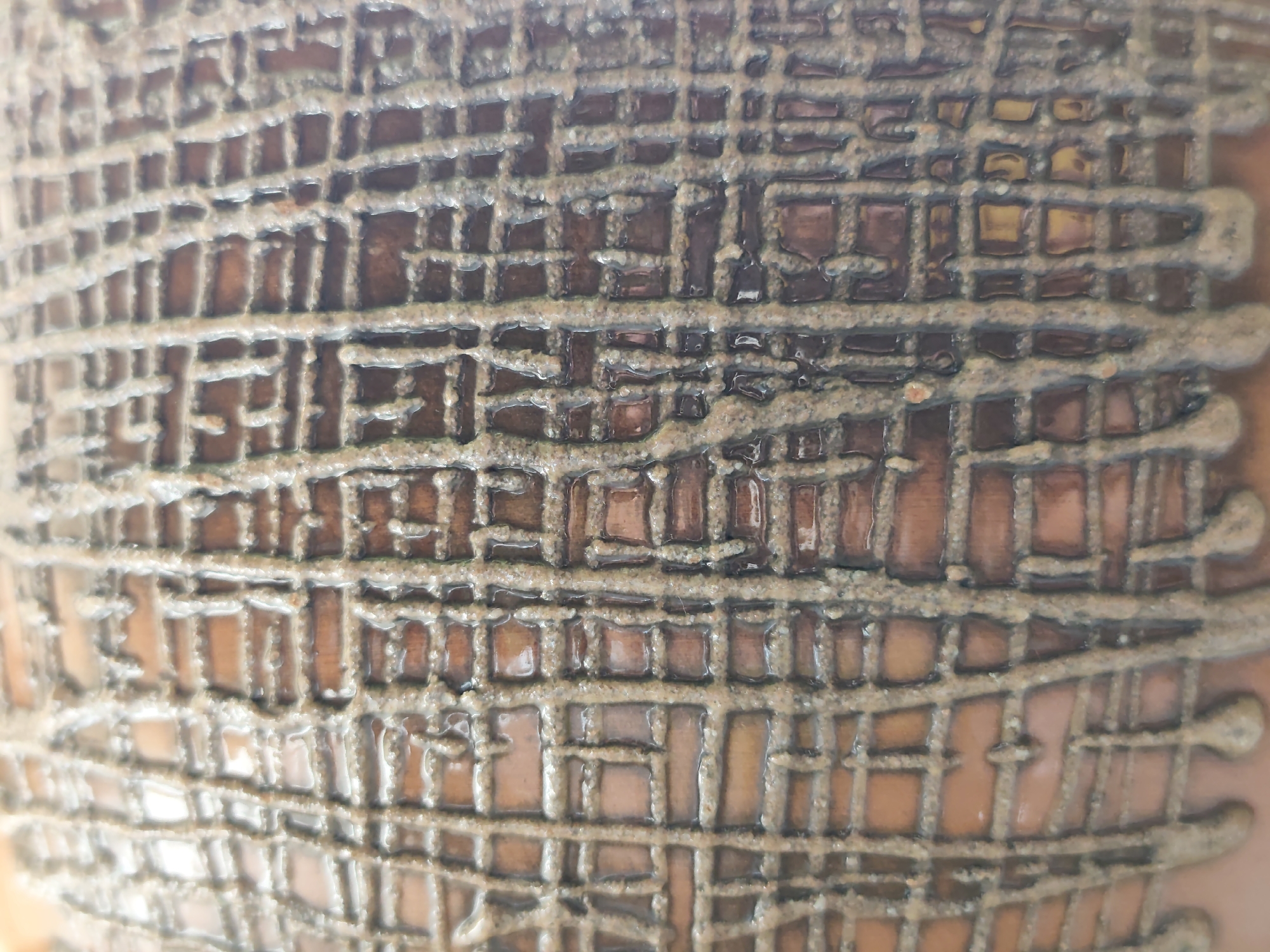 The image displays a close-up view of a textured ceramic surface. The surface is adorned with an intricate, grid-like pattern formed by raised lines of a lighter, granular material against a darker, smooth background. Within the grid, the darker background is divided into numerous rectangular and square compartments, some of which appear to be filled with a glossy, amber-colored substance, while others are open and reveal the underlying texture. The overall impression is of a decorative or functional object with a deliberate, handcrafted design. There is no discernible location in the city of Minsk, Belarus, nor are there any people, actions, or interactions depicted in this abstract surface detail.