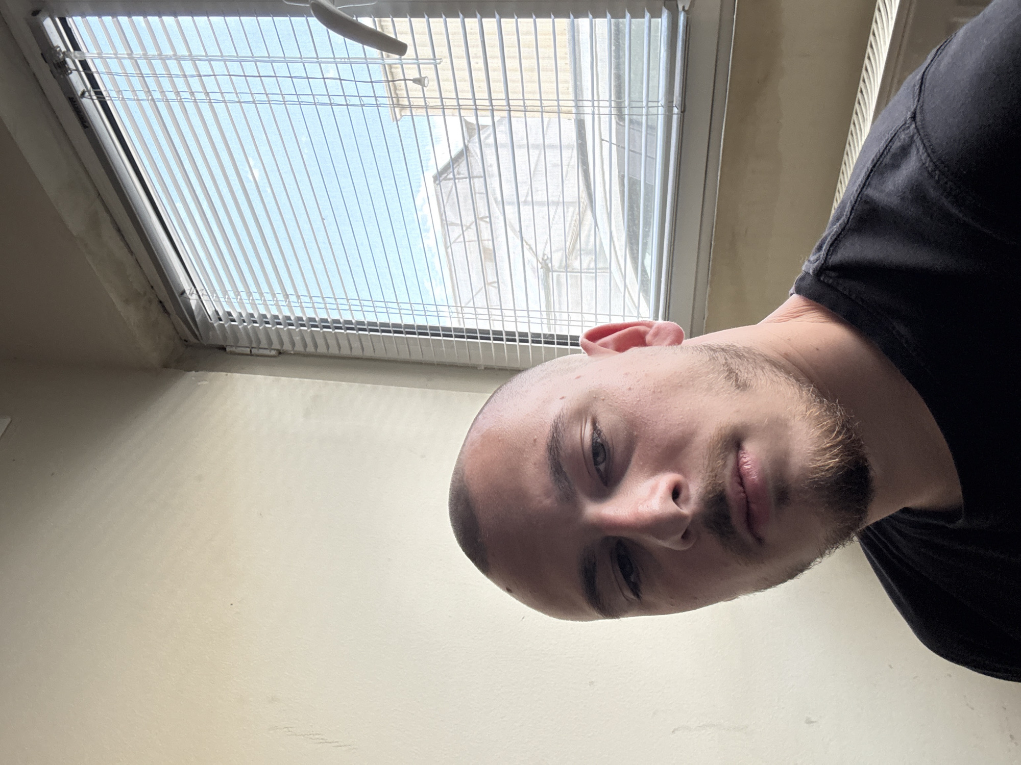 The image is a close-up selfie of a bald man with a short beard. He is wearing a black shirt. The background shows a window with white blinds. Outside the window, a structure of some kind is visible, possibly a building or scaffolding. The wall adjacent to the window is painted light beige. There is no identifiable information to locate the image at a precise location within the city of Stara Zagora, Bulgaria.