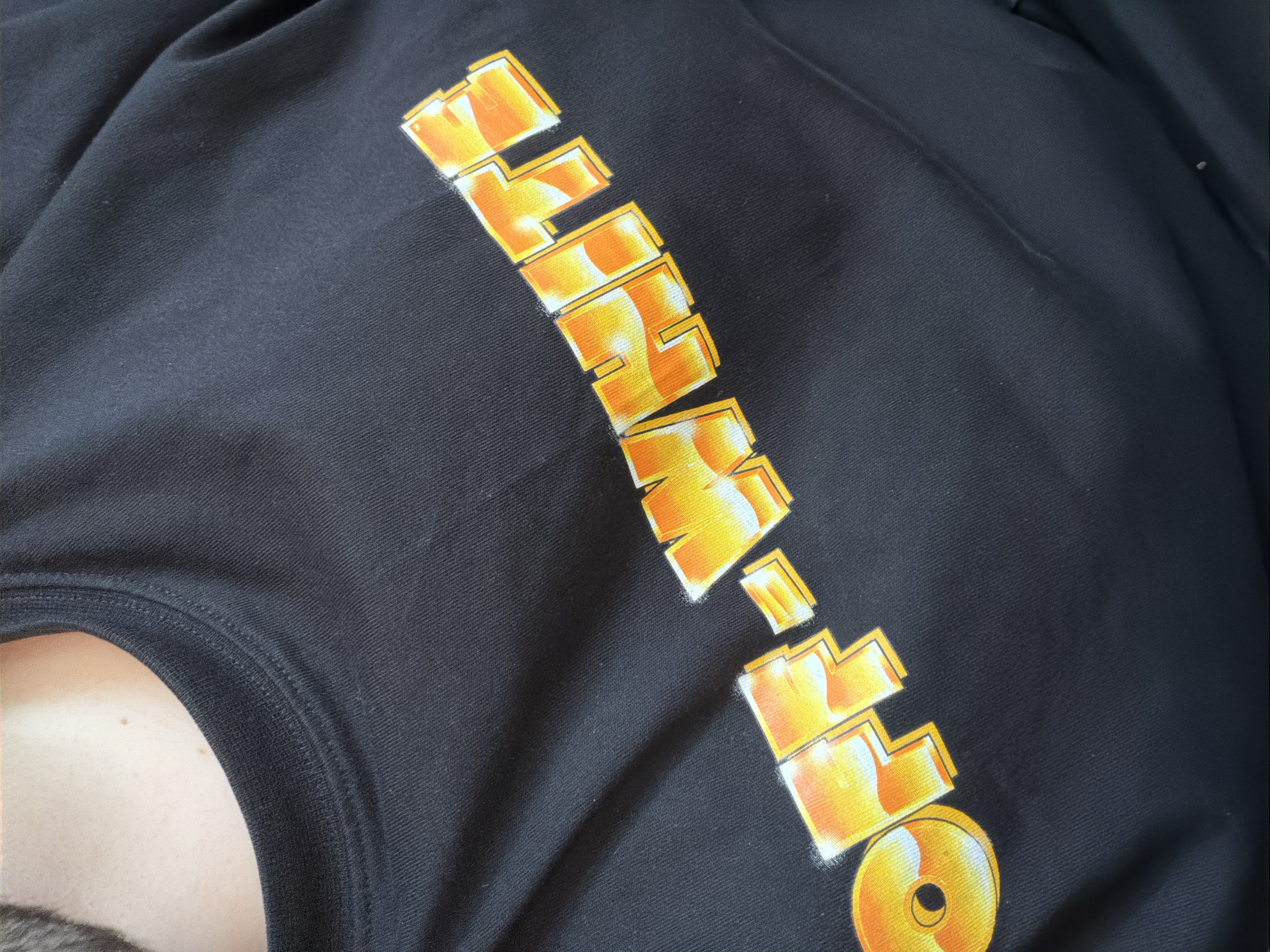 This close-up photograph, taken indoors in Sofia, Bulgaria, features a person wearing a black garment, likely a t-shirt or sweatshirt. The primary subject is a large graphic print emblazoned across the chest of the garment. The print spells "OFF-WHITE" in stylized, bold block letters, angled across the fabric. The text is rendered in vibrant shades of orange and yellow, with distinct white highlights and dark outlines that create a three-dimensional effect. The final letter "O" is notably designed as a complete circle with a smaller inner circle, resembling a disc or target.
The black fabric of the garment has a visible fine ribbed texture. Some minor lint or dust particles are present on the fabric surface. A portion of the wearer's body is visible in the bottom left of the frame, including their fair skin at the neck and upper chest, a small mole, and the edge of some facial hair. The ribbed collar of the garment is also clearly seen around the neckline. The lighting is soft and even, suggesting an indoor environment without harsh direct light. No specific activity is depicted, and the person's face is not shown.