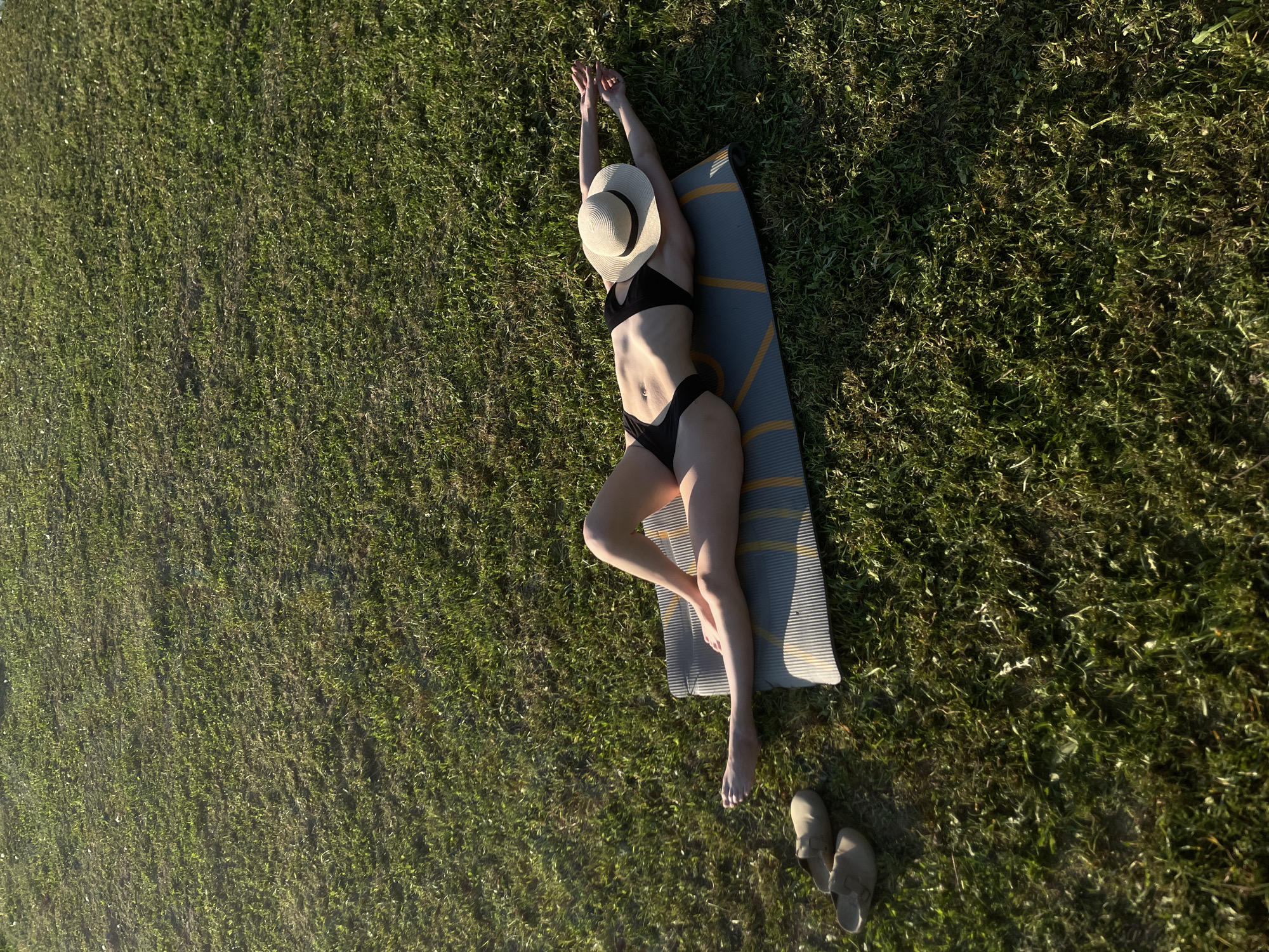 A woman in a black bikini and a straw hat is lying on a yoga mat in a grassy area. Her arms are raised above her head. A pair of slippers are located near the bottom right of the frame. The photo appears to have been taken from directly above, looking down. It is possible that the location is in the city of Подшивалово, Russia.