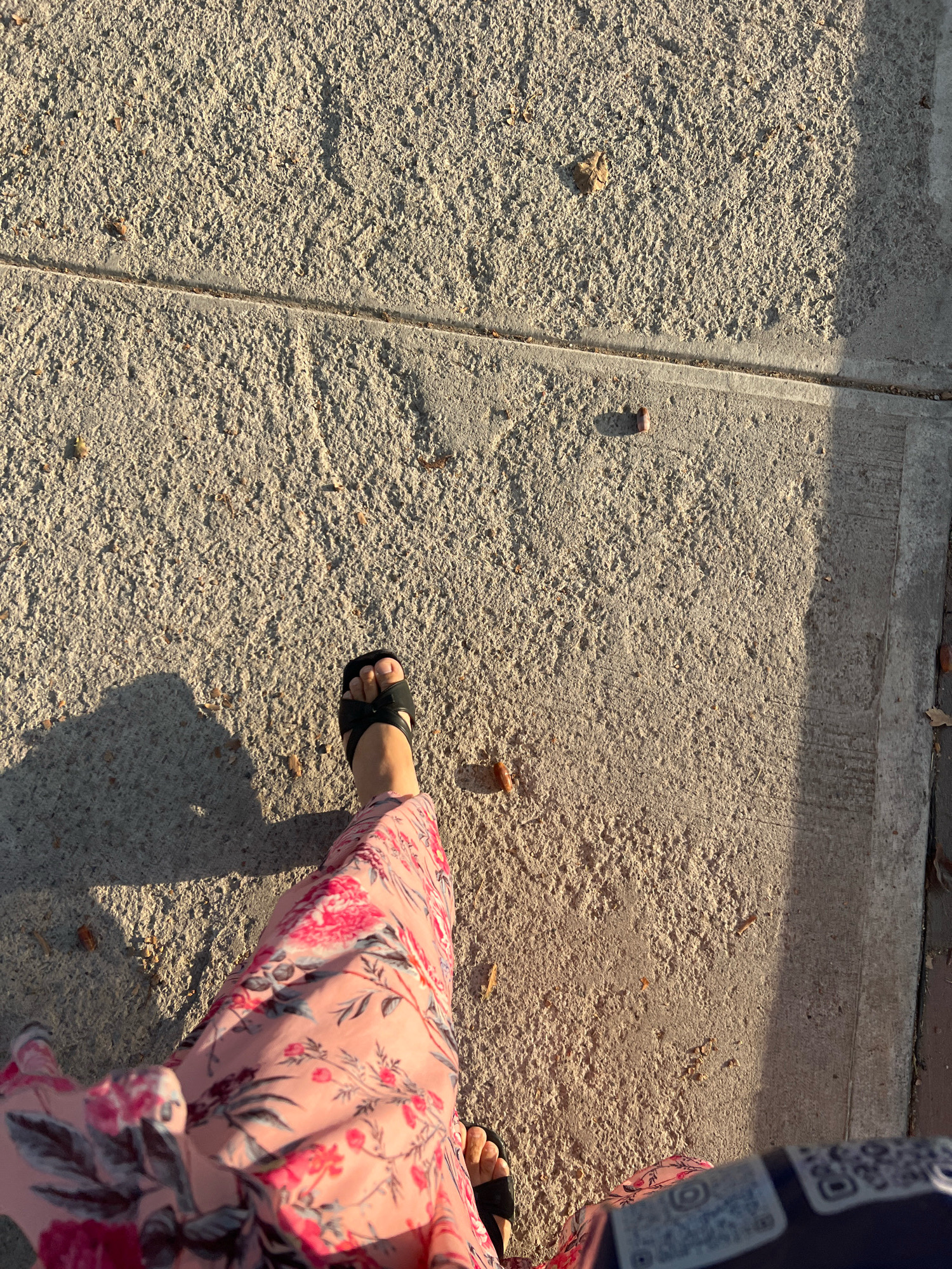 A person's legs and feet are shown from a high-angle perspective, walking on a textured concrete surface. The person is wearing a flowing pink floral-patterned maxi skirt and black open-toed sandals. A shadow of the person is cast on the ground to the left. The concrete appears to be part of a sidewalk or walkway, with a visible expansion joint running horizontally across the frame. Small debris, possibly leaves or twigs, are scattered on the concrete. The scene is illuminated by sunlight, creating strong shadows and highlighting the texture of the ground. The lower right corner of the image shows a portion of a dark blue object with white text and QR codes.