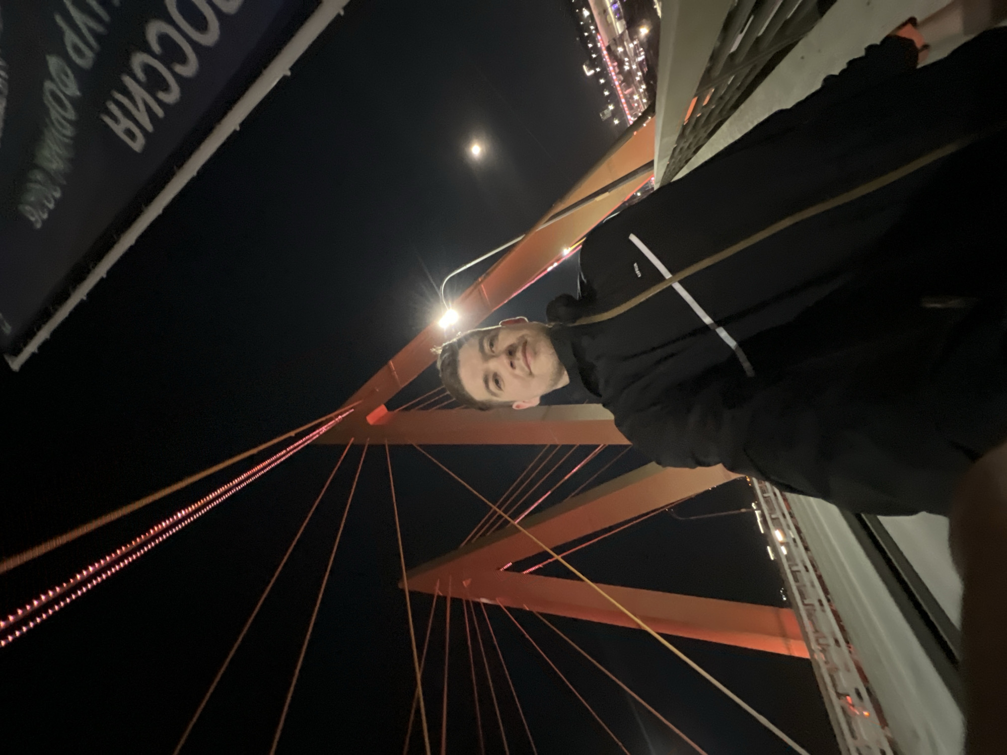 A man is on the Millennium Bridge in Kazan, Russia at night. He is facing the camera. The bridge's red cables are visible in the background, along with the night sky and moon. The Russian word "РОССИЯ" is visible on a sign.