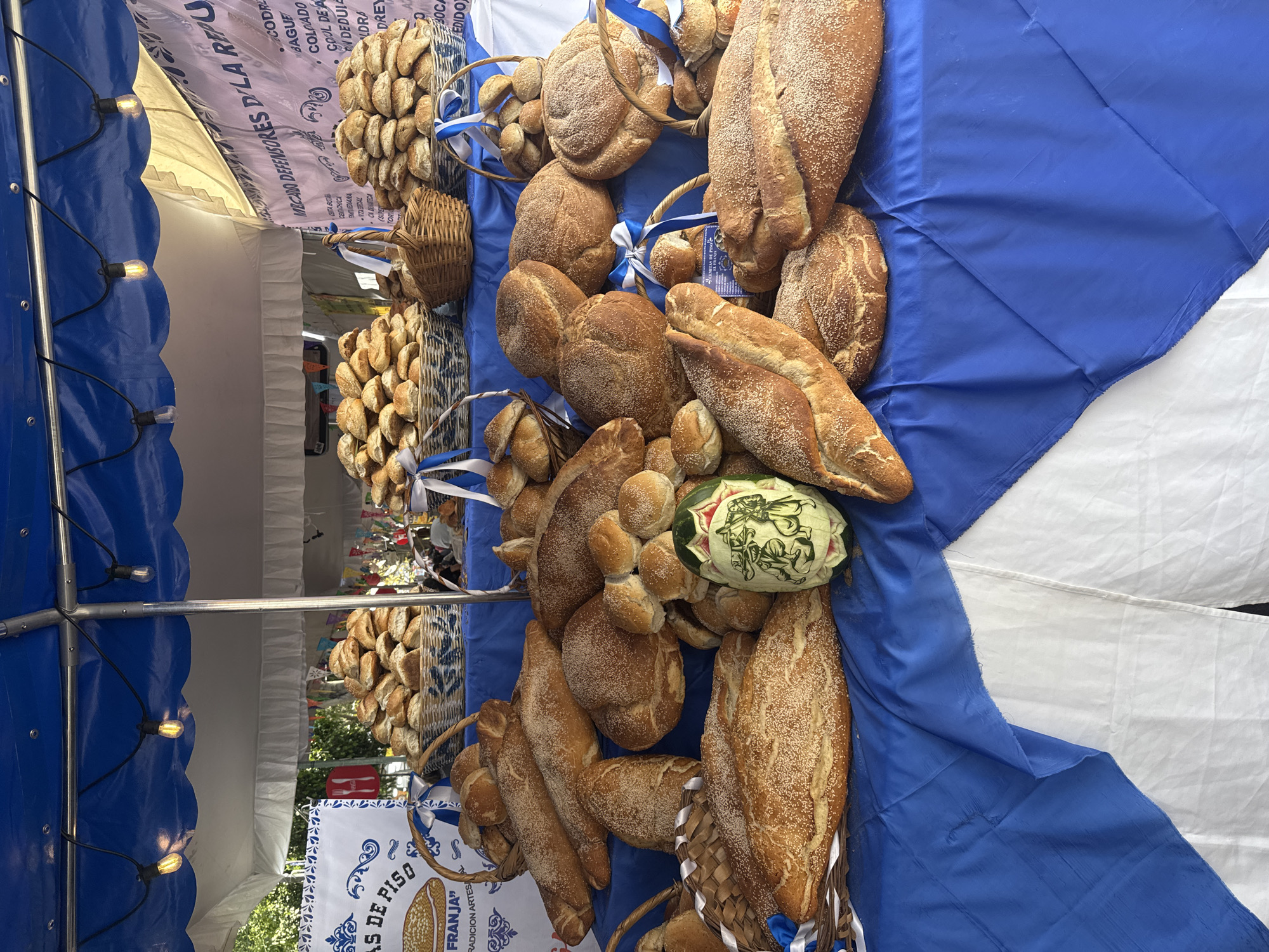 A collection of various types of bread, including loaves and rolls, are displayed on a blue and white cloth. A decorated melon sits among the bread. The bread is in baskets adorned with blue and white ribbons. In the background, stacks of small breads are visible. The scene appears to be a market stall, possibly in Puebla, Mexico, judging by the sign referring to "Defensores de la Republica".