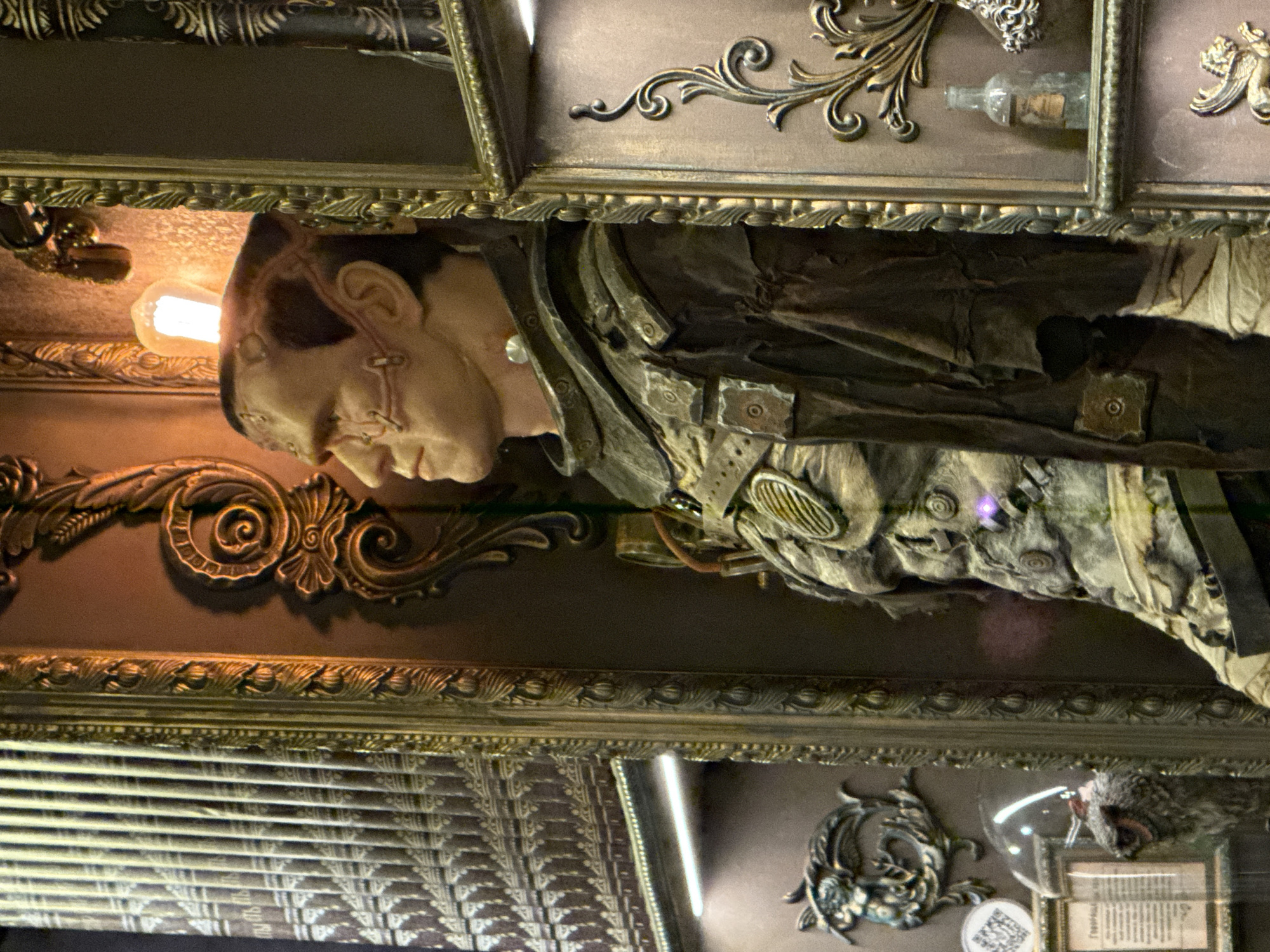 A sideways close-up shot depicts a detailed, sculptural representation of Frankenstein's monster enclosed within a gilded, ornate display case. The case features intricate filigree and scrollwork designs in a metallic hue. Inside, the monster's face, turned in profile, showcases stitches and a bolt on the neck. The creature is dressed in worn, layered garments with metallic accents. An exposed lightbulb illuminates the scene. The background reveals additional decorative elements within the case, including a bottle and antique accessories. The location of the scene is Moscow, Russia.