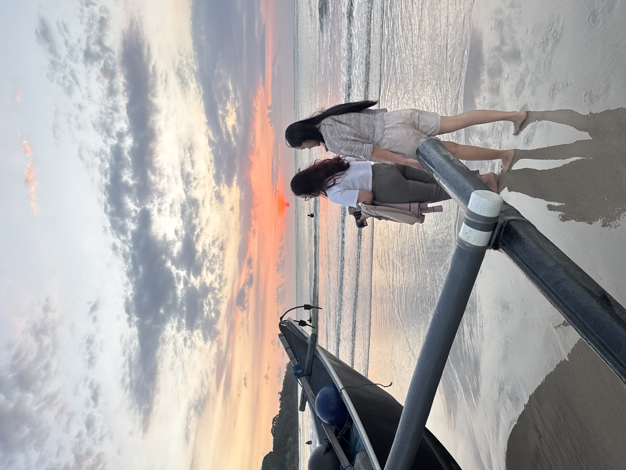 The image captures a serene sunset scene on a beach in Kuta Selatan, Indonesia. In the foreground, partially obscuring the view on the right, is a section of a small, dark-colored boat, likely a fishing boat or a traditional Indonesian jukung, with two grey poles angled across the frame. A blue buoy and some ropes are visible within the boat.
Two women stand barefoot on the wet sand, facing the ocean and the setting sun. The woman closer to the foreground has long, dark hair, wears a white t-shirt, dark trousers, and glasses, and appears to be holding a smartphone or a similar device. The other woman, positioned slightly behind and to her left, also has long, dark hair and is dressed in a light-colored, patterned top and light shorts. Both women's reflections are visible in the highly reflective, wet sand beneath them.
The sky is ablaze with the warm, vibrant hues of sunset, transitioning from bright orange and pink near the horizon to softer grey and white clouds higher up. These dramatic sky colors are mirrored in the calm ocean water, where gentle waves lap the shore, creating reflective patterns. The overall atmosphere is peaceful and picturesque, highlighting the beauty of the tropical twilight. No visible text is present in the image.