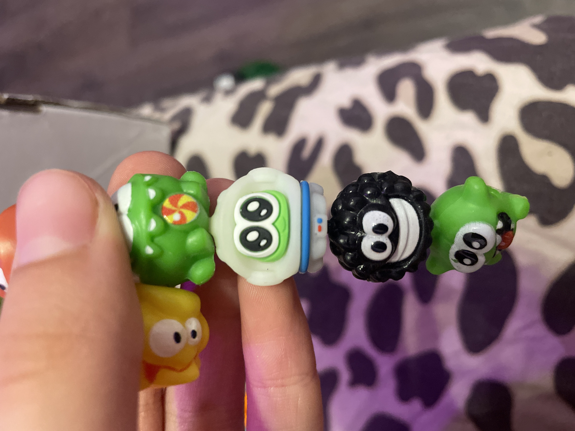 A hand is holding a stack of six small, colorful, toy figures. The figures are arranged vertically and appear to be made of plastic. The topmost figure is partially obscured by a thumb and is red and orange. Below that is a yellow figure with two large white eyes. Next is a green figure with a candy-like design on its chest. The fourth figure is white and resembles an astronaut with large green-outlined eyes and a blue band around its helmet. The fifth figure is black and fuzzy with large white-eyed black pupils and a wide smile. The bottommost figure is green and has large eyes and an open mouth with a red tongue. The background consists of a patterned fabric, possibly a blanket or upholstery, with irregular dark shapes on a lighter background. There is no indication of the city of Kokkorevo, Russia, in the image.