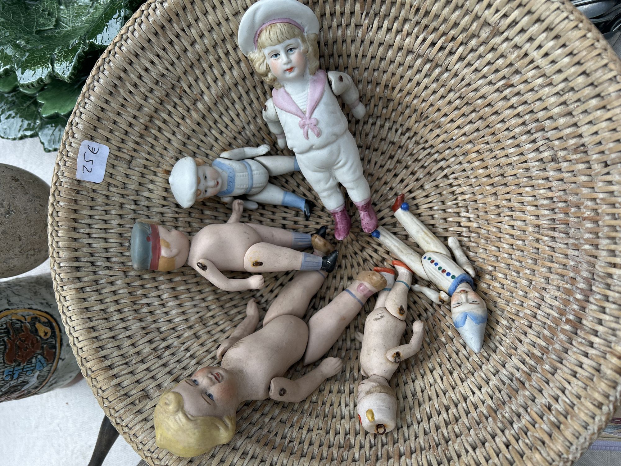 The image shows a collection of antique dolls in a wicker basket. There are five dolls visible, each with different clothing and features. One doll is dressed in a white sailor suit with pink accents, another in a blue and white sailor outfit, and the others are nude or partially dressed. The dolls appear to be porcelain or bisque. The basket is woven and has a natural color. A price tag of "€352" is visible near the left edge of the basket. The background is slightly out of focus, but includes some greenery and other items suggesting the basket is on a table or display area. The setting is possibly an antiques market or store, potentially in Paris, France, given the Euro currency symbol and the style of the antiques.