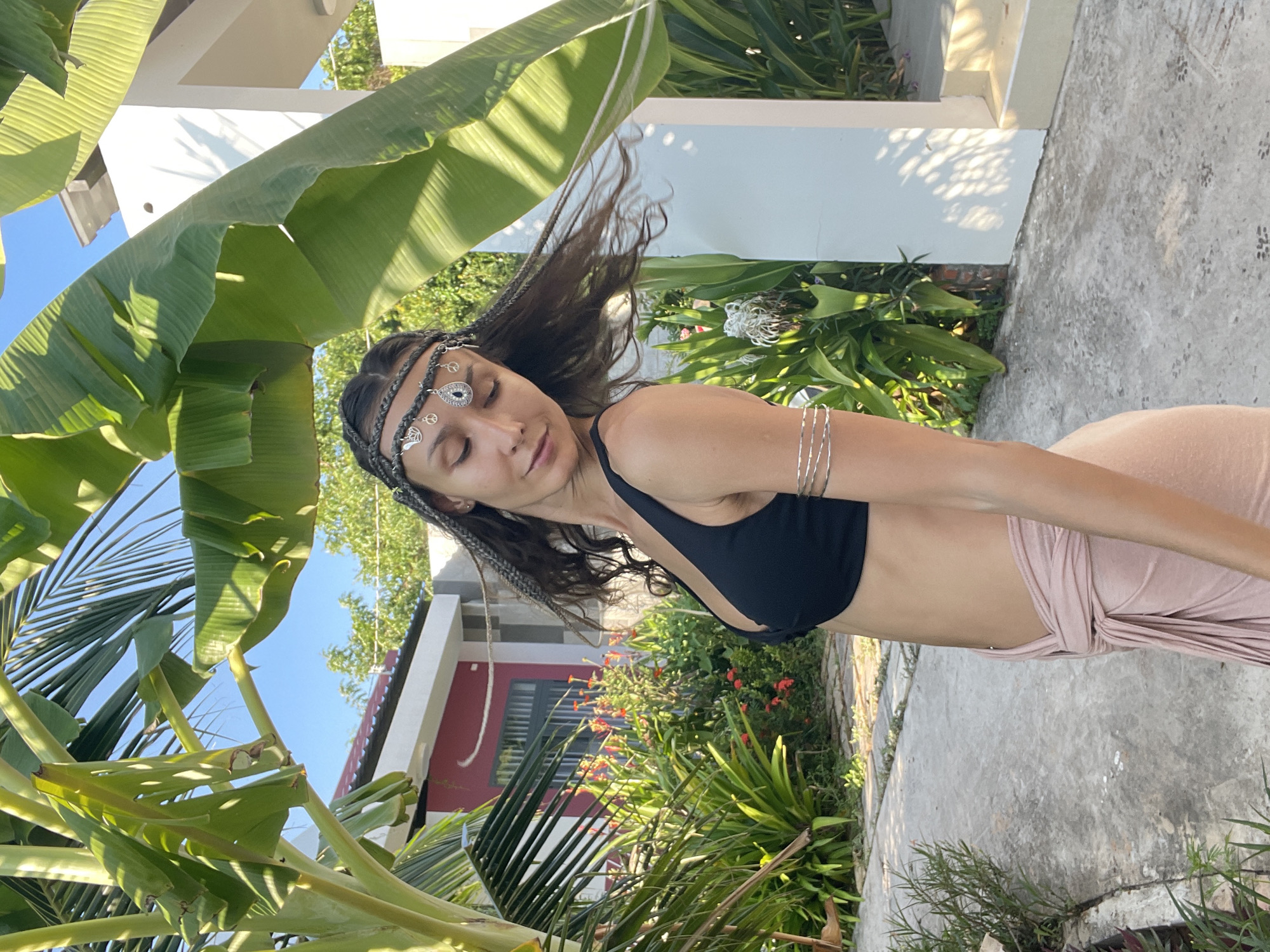 In Mũi Né, Vietnam, under a clear blue sky, a woman stands outdoors, posing with her head tilted down and her eyes closed. She has her hair in braids and is adorned with a decorative headband featuring various charms, including a teardrop-shaped pendant and peace signs. She is wearing a black sports bra and a light pink wrap skirt, tied at her waist. Multiple thin silver bracelets adorn her upper arm.
The woman is positioned in front of a lush garden with vibrant green plants and red flowers. Behind the garden, a building with a red and white facade is visible, along with parts of a white structure that appears to be a porch or walkway. Sunlight casts dappled shadows on the ground and the white structure, suggesting it is mid-day. The ground is paved with concrete tiles. Large banana leaves and palm fronds frame the scene from above, creating a tropical atmosphere.