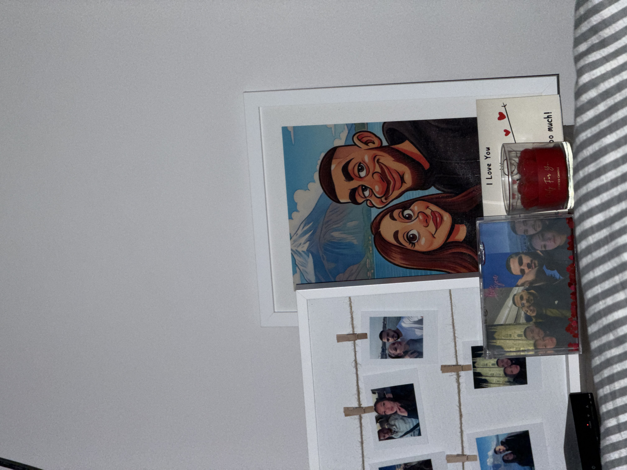 A collection of personal items is displayed indoors in Varna, Bulgaria, against a light gray wall. On the left, a white-framed board holds multiple small, square-format photographic prints suspended by twine and miniature wooden clothespins. These prints show various individuals, including a man and woman in a car, a couple embracing outdoors, a couple in a scenic outdoor setting, and a couple positioned in front of an architectural landmark resembling Milan Cathedral. To the right, a larger white-framed print features a stylized cartoon portrait of a smiling man with short dark hair and a beard, alongside a smiling woman with long reddish-brown hair, set against a background of mountains and a body of water.
In the foreground, from left to right, a portion of a black electronic device with a red indicator light is visible on a striped gray and white fabric surface. Adjacent to this, a transparent rectangular display case contains a group photograph of multiple individuals and features red decorative elements at its base, with "I love you" text partially visible. To its right, a clear glass container holds a red candle labeled "Only For You. SCENTED CANDLE." Beside the candle, a white card with red hearts is present, displaying the text "I Love You" and "too much!"