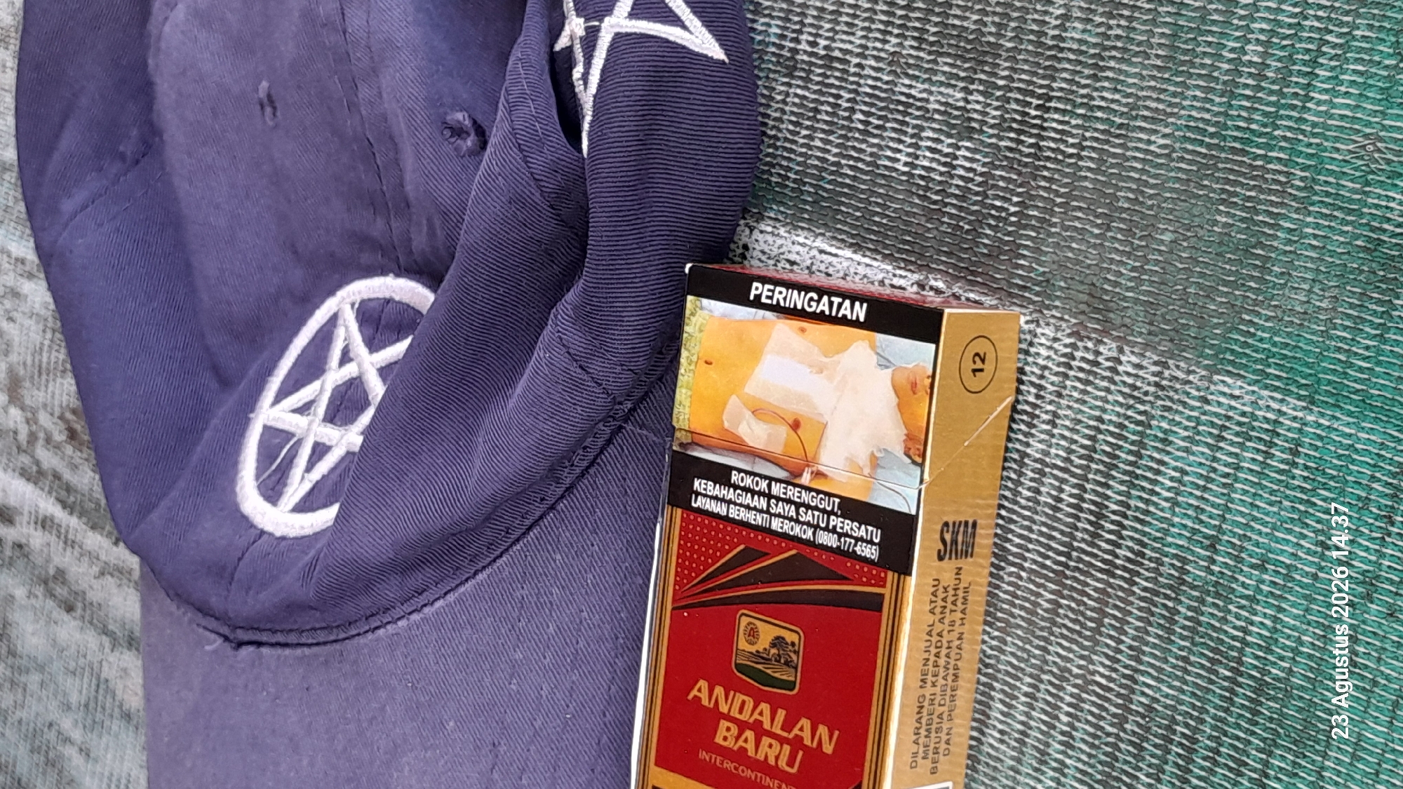 A blue baseball cap with a white embroidered pentagram is resting on a textured, greyish-green surface. In front of the cap, a red and gold cigarette pack, labeled "ANDALAN BARU," is positioned diagonally. The cigarette pack features Indonesian text and a warning graphic of a young boy coughing. A circular "12" is visible on the lower right side of the pack. A timestamp reading "23 Agustus 2026 14:37" is in the bottom left corner of the image. The scene is indoors or shaded, with no visible natural light source. There are no people or animals present in the image. The location is identified as Gemaharjo, Indonesia.