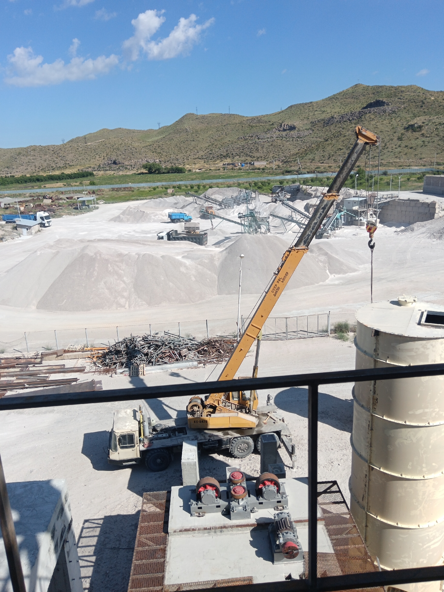 This media file captures an outdoor industrial scene, likely a quarry or aggregate processing plant, under a bright, sunny sky with scattered white clouds. The terrain is mountainous and arid, with dry, green vegetation on the slopes. The setting appears to be Aragatsavan, Armenia, as indicated by the location context.
The primary subjects are heavy machinery, including a large yellow crane with its boom extended, a mobile crane, and several trucks. Piles of white aggregate or sand dominate the landscape, indicating a site for material extraction and processing. In the foreground, there are some metal beams and a large pile of scrap metal. Also visible are large cylindrical storage silos.
The crane appears to be in operation, with its hook suspended over one of the silos, suggesting an ongoing task of lifting or moving materials. The mobile crane is parked nearby. The presence of trucks implies the transportation of materials to or from the site.
The time of day is clearly daytime, likely mid-morning to afternoon, given the strong shadows cast by the machinery and the bright sunlight. The weather is fair and clear. There is no visible text in the image, except for what appears to be a license plate on the mobile crane, but it is too small and blurry to read. The overall impression is one of industrial activity in a rural, mountainous environment.
