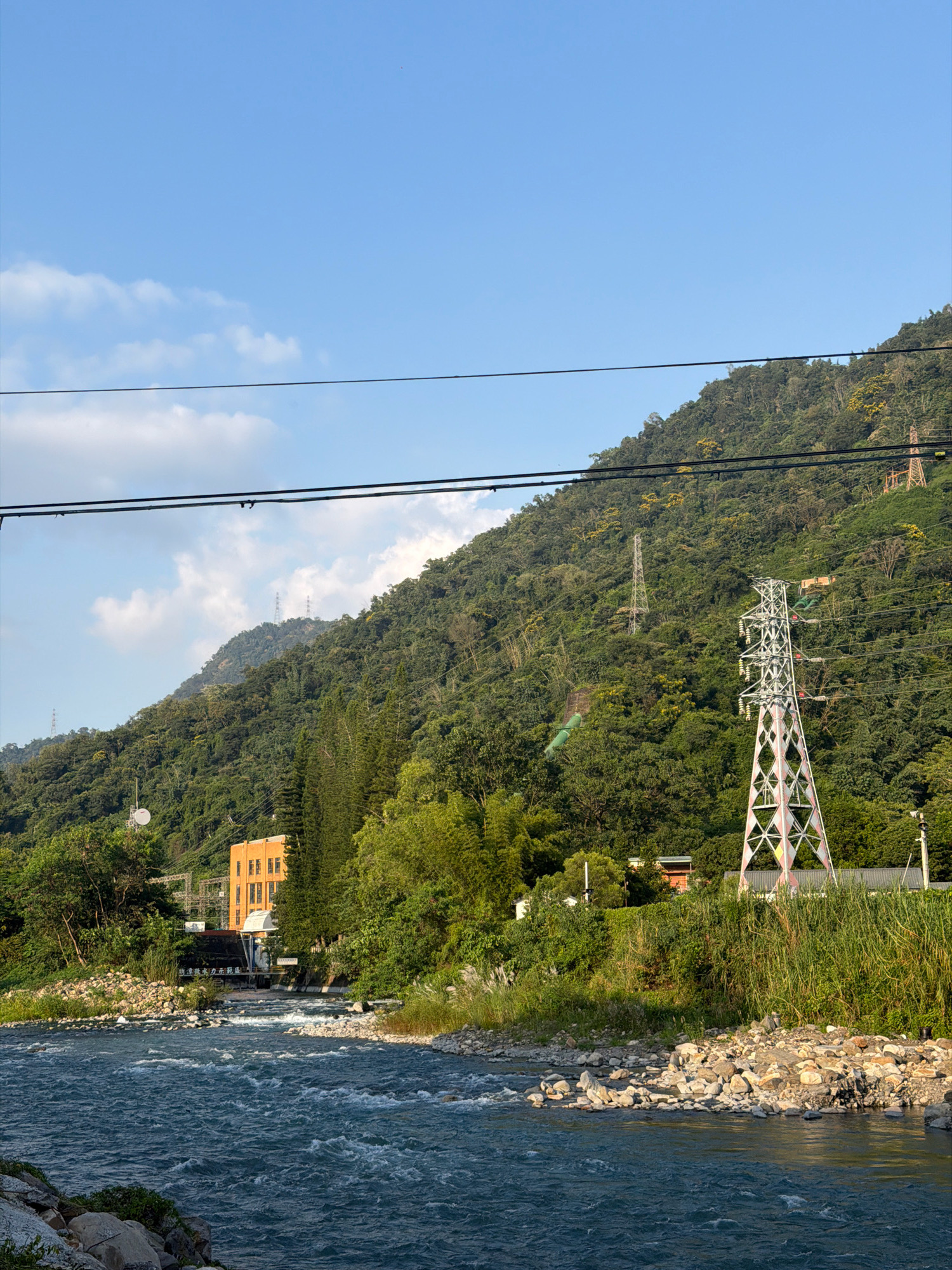 This image depicts a scenic view of the Shuili River in Shuili Township, Taiwan. The river, with its characteristic blue-green water and rocky banks, flows through the foreground. Lush, green mountains covered in dense foliage dominate the background, with several power transmission towers visible on their slopes.
On the left side of the image, a yellow brick building, likely a power station, is situated near the riverbank. A white satellite dish is mounted on a pole in front of the building. Tall, dark green coniferous trees stand to the right of the building, partially obscuring it.
The middle ground features a vibrant green landscape with various trees and vegetation. In the right portion of the image, a tall, white and pink lattice power transmission tower stands prominently. A smaller, more distant transmission tower can be seen further up the mountain. The sky above is a clear blue with a few scattered white clouds.
The overall scene captures the natural beauty of the mountainous region, interspersed with infrastructure related to power generation and transmission. The image is a still, unposed shot, likely taken during daylight hours. No people are visible in the frame.