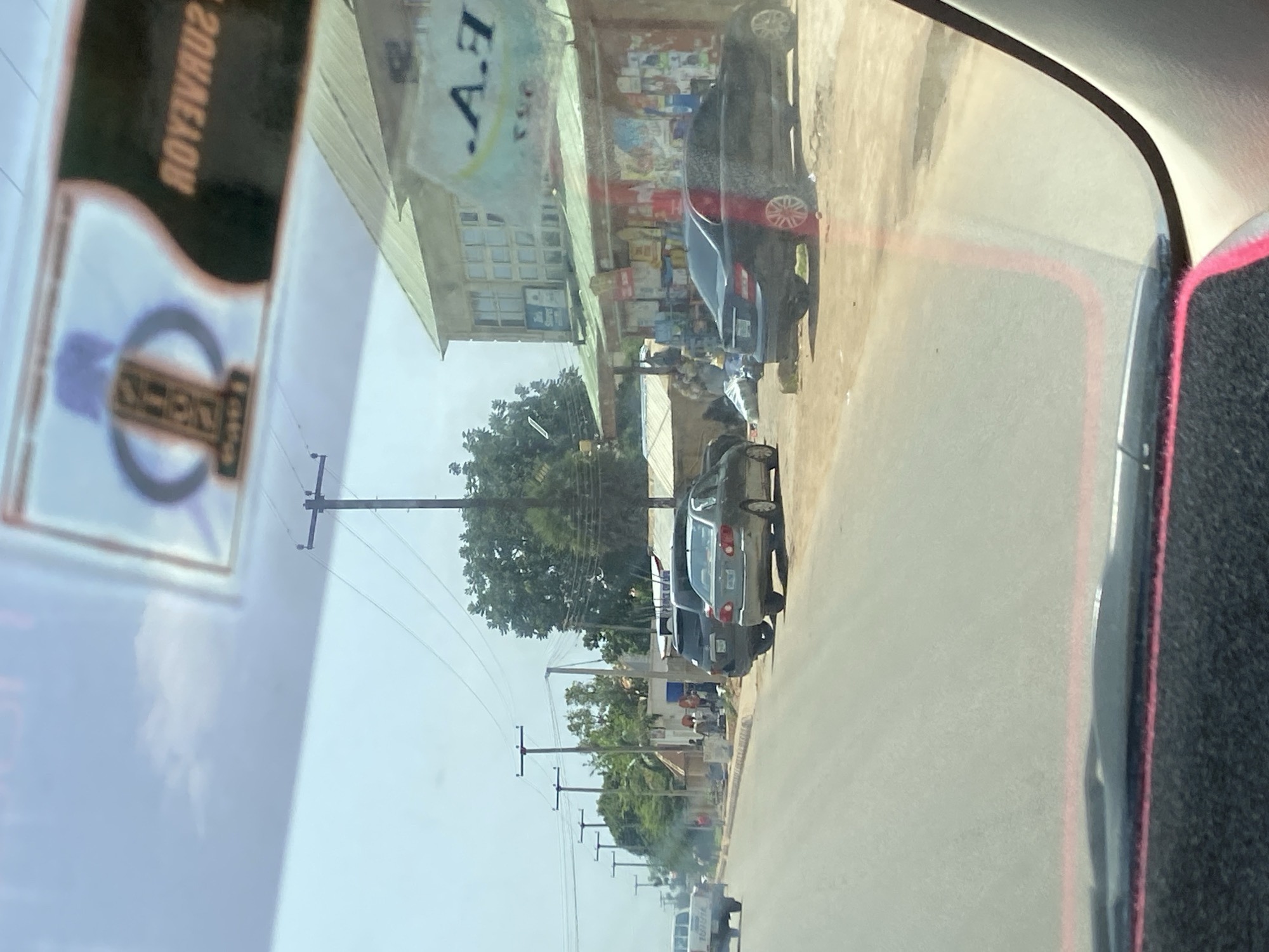 The image depicts a street scene in Bar Jirgi Summa, Nigeria, viewed from inside a vehicle. The foreground shows the interior of a car, with a black dashboard and a pink trim visible along the bottom edge. The windshield is slightly dirty, and a sticker with what appears to be text and a circular emblem is affixed to the upper part.
Beyond the windshield, the street is paved and slopes slightly downhill from left to right. Several vehicles are parked along the right side of the road, including a silver sedan, a dark SUV, and a black sedan. Further down the street, a white pickup truck is seen moving away from the viewer.
On the right side of the street, there are several buildings and structures. One building has a green, sloped roof and appears to be a shop or commercial establishment with visible signage. Goods are displayed outside this building. Other structures, possibly stalls or less permanent shops, are also present, with various items including what look like sacks and other merchandise. Power lines and utility poles are visible throughout the scene, running along the side of the road and over the buildings. A cluster of trees and dense foliage is present in the background, suggesting a semi-urban or peri-urban environment. Several individuals are visible on the side of the road, some near the buildings and others further down. The sky is overcast with a few clouds visible.