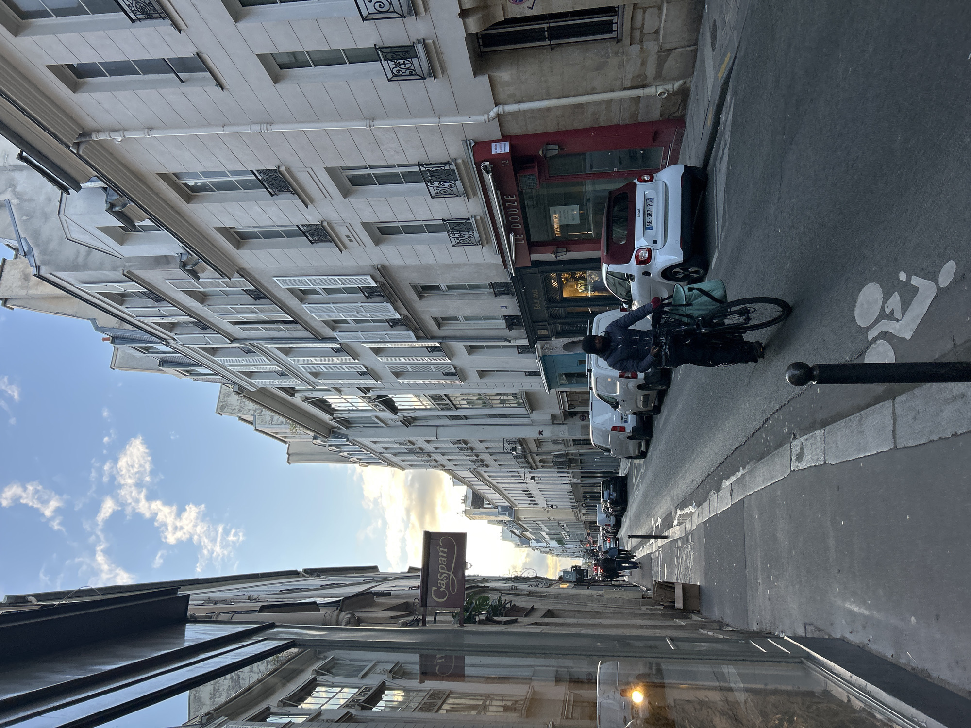 The image captures a narrow street in an urban setting, likely Paris, France. Buildings with multiple stories line both sides of the street, displaying classic architectural features like rows of windows with small balconies. The sky above is partially visible with scattered clouds.
A man is on a bicycle, positioned on the right side of the street. Several parked cars line the same side of the street. On the left side, a sign that reads "Caspari" is visible. The foreground shows elements of a storefront, including a window. The overall light suggests it is either late afternoon or early morning.